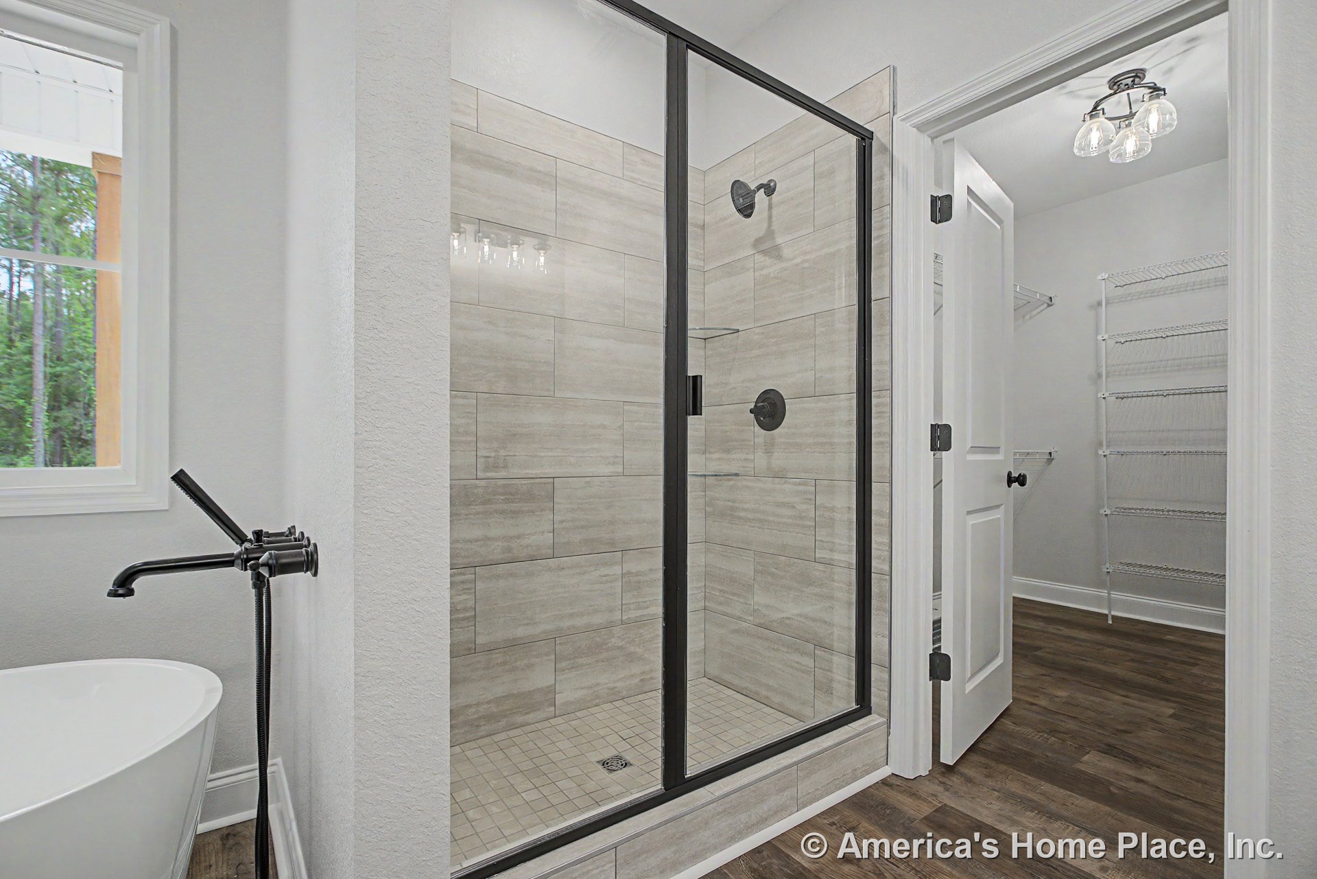 Walk-in shower with frameless glass door and large beige rectangular tile, freestanding bathtub with black faucet, wood-look flooring, walk-in closet with wire shelving, white trim
