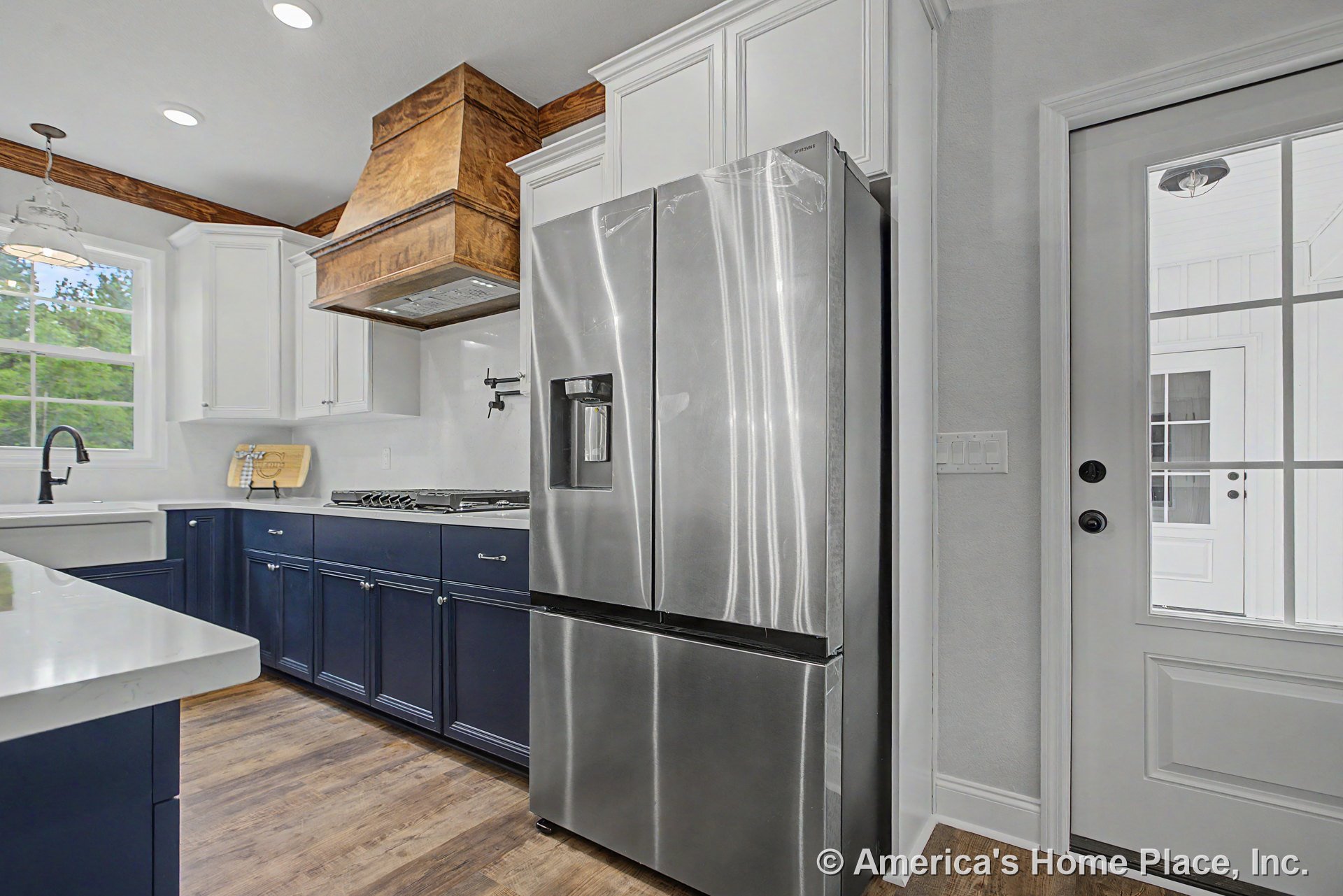 Navy base cabinets and white upper cabinets with crown molding, stainless steel refrigerator, wood vent hood above cooktop with pot filler, farmhouse sink beneath window, white