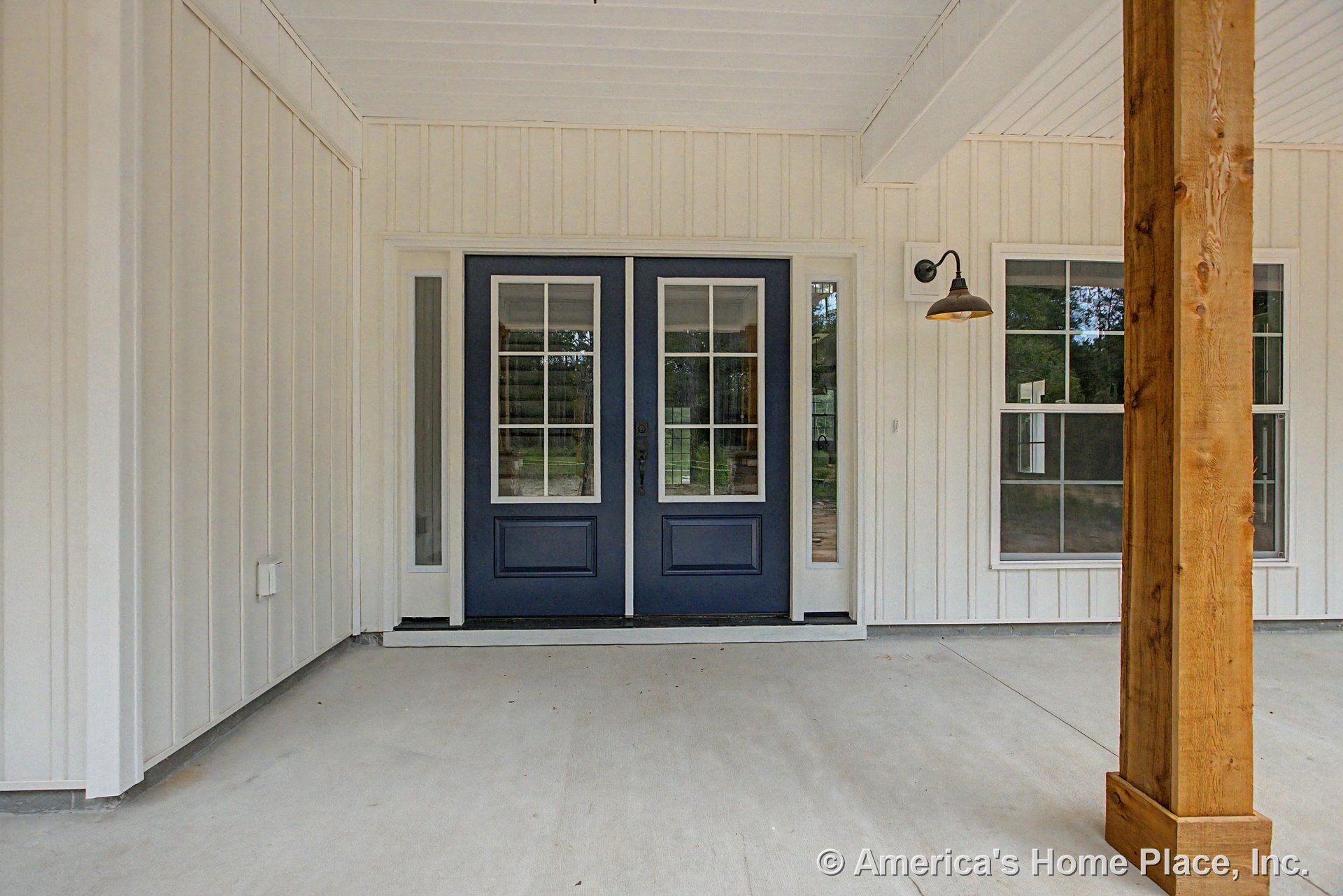 Double blue entry doors with glass panels and transom windows, vertical board and batten siding, covered porch ceiling, concrete floor, exterior wall-mounted light fixture, and