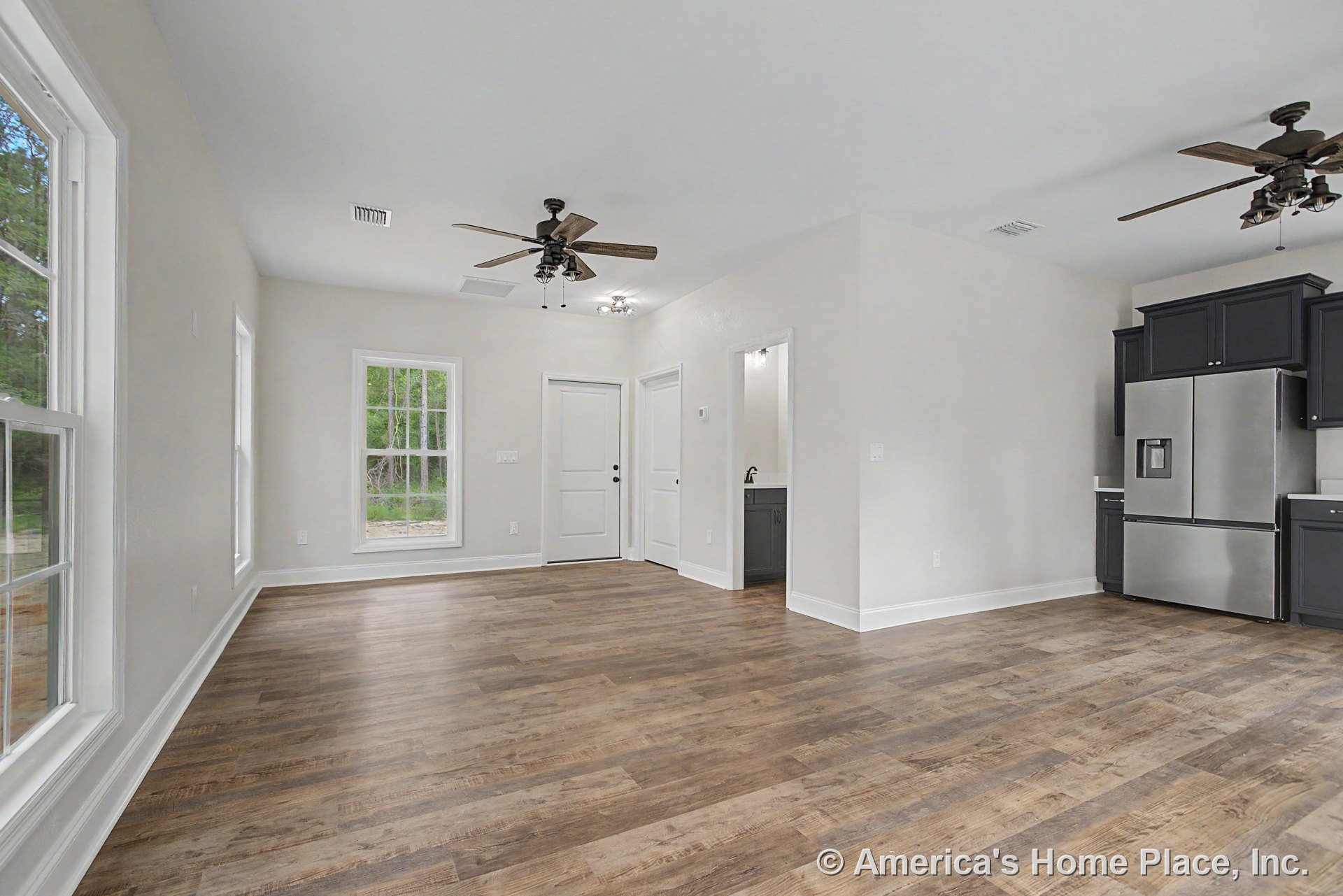 Open floor plan with wood-look plank flooring, large double-hung windows, dark kitchen cabinets, stainless steel refrigerator, ceiling fans with lights, white trim and baseboards.