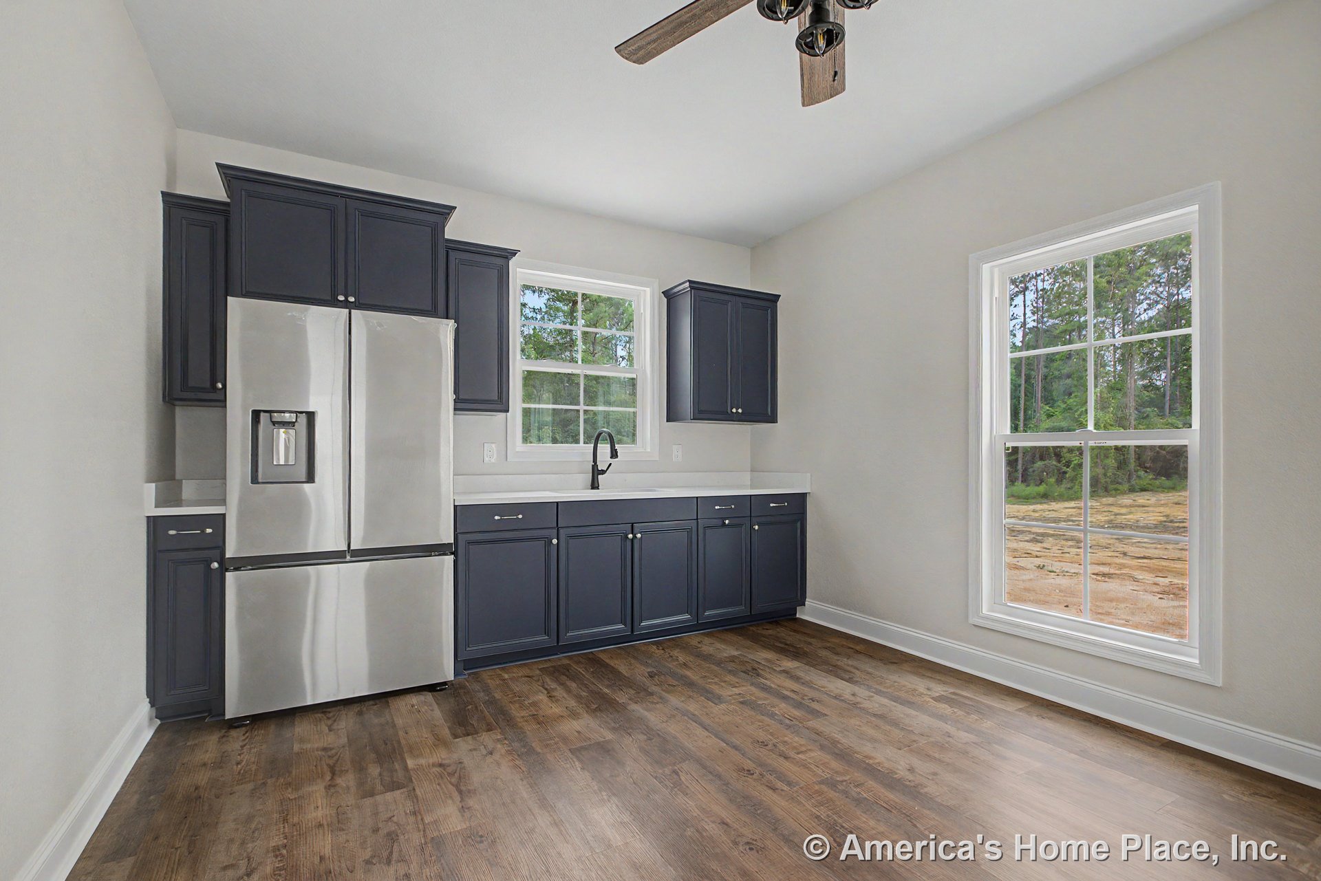 Dark upper and lower cabinets with stainless steel refrigerator, white solid surface countertops, single bowl sink, double-hung windows, wood-look vinyl plank flooring, ceiling fan