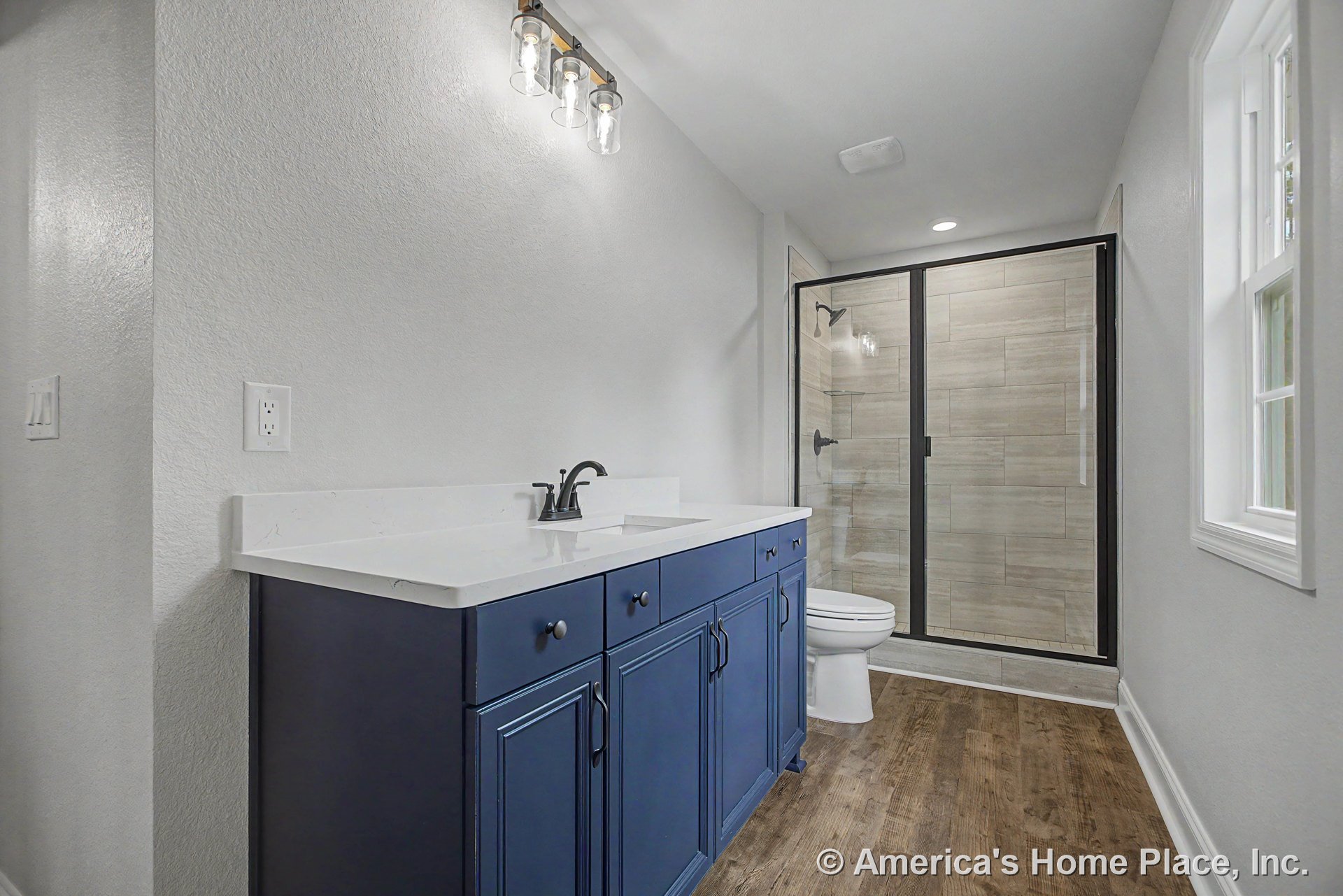 Blue bathroom vanity with white solid surface countertop, glass shower enclosure, wood-look vinyl flooring, wall-mounted light fixture, double-hung window with white trim, and