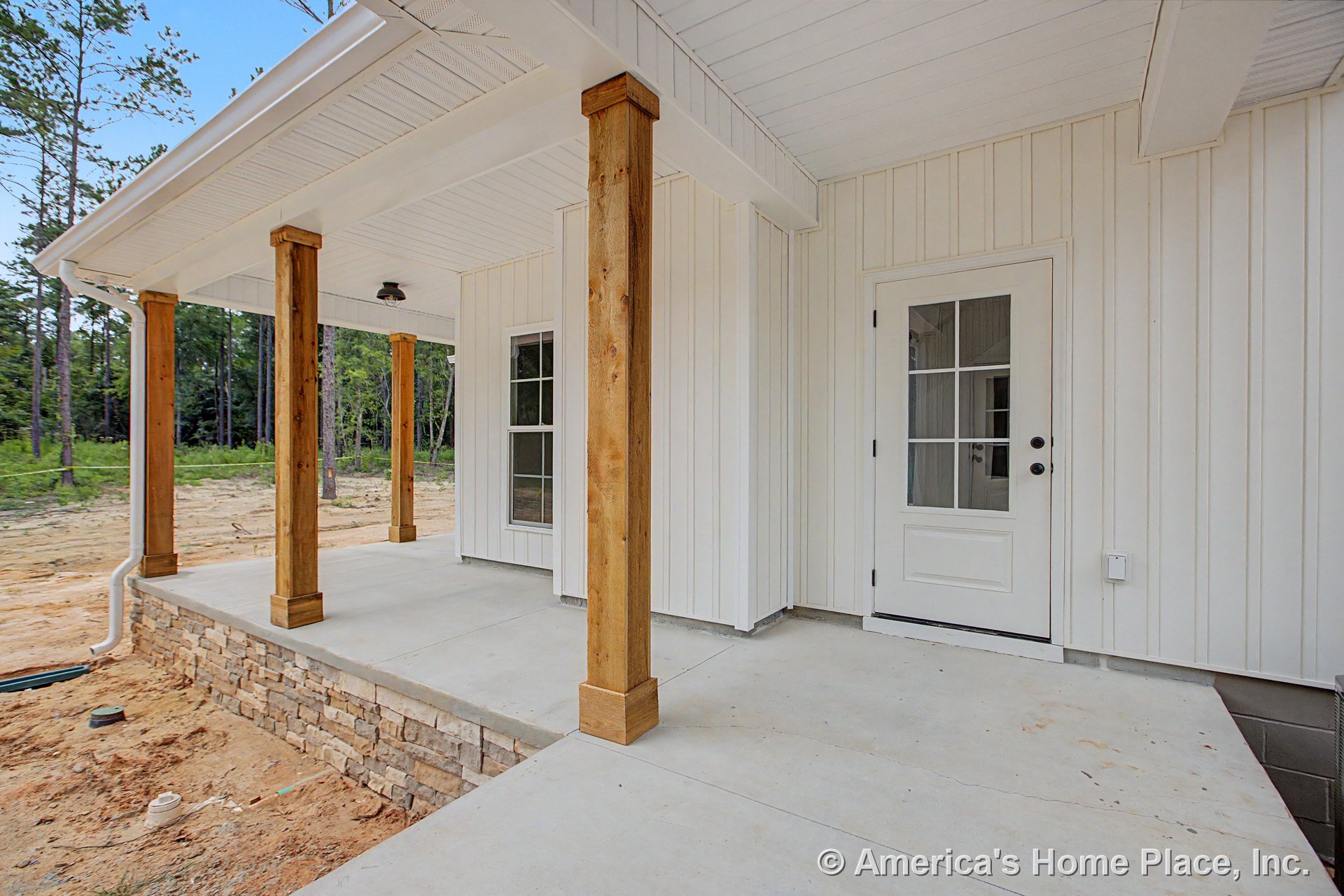 Covered front porch with wood support columns, stone skirting along the foundation, vertical white siding, white exterior door featuring glass panes, concrete porch floor, outdoor