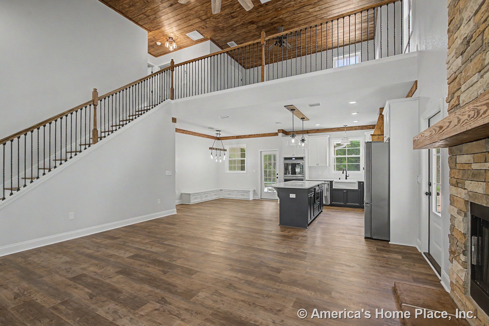 Vaulted wood ceiling and window wall above open-concept living area with stone fireplace and wood mantel, adjacent to modern kitchen featuring a large island with pendant lighting