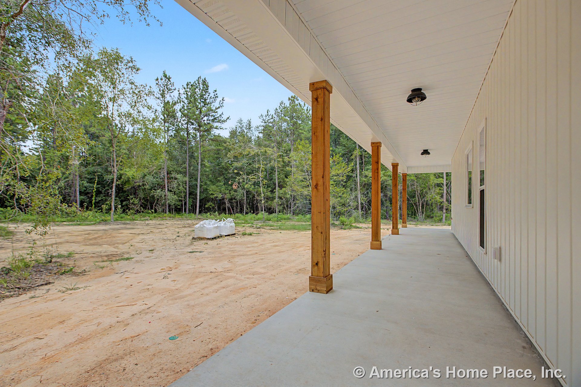 Covered front porch with exposed wood support columns, concrete flooring, white vertical vinyl siding, double window units with trim, overhead lighting fixture, and vinyl soffit
