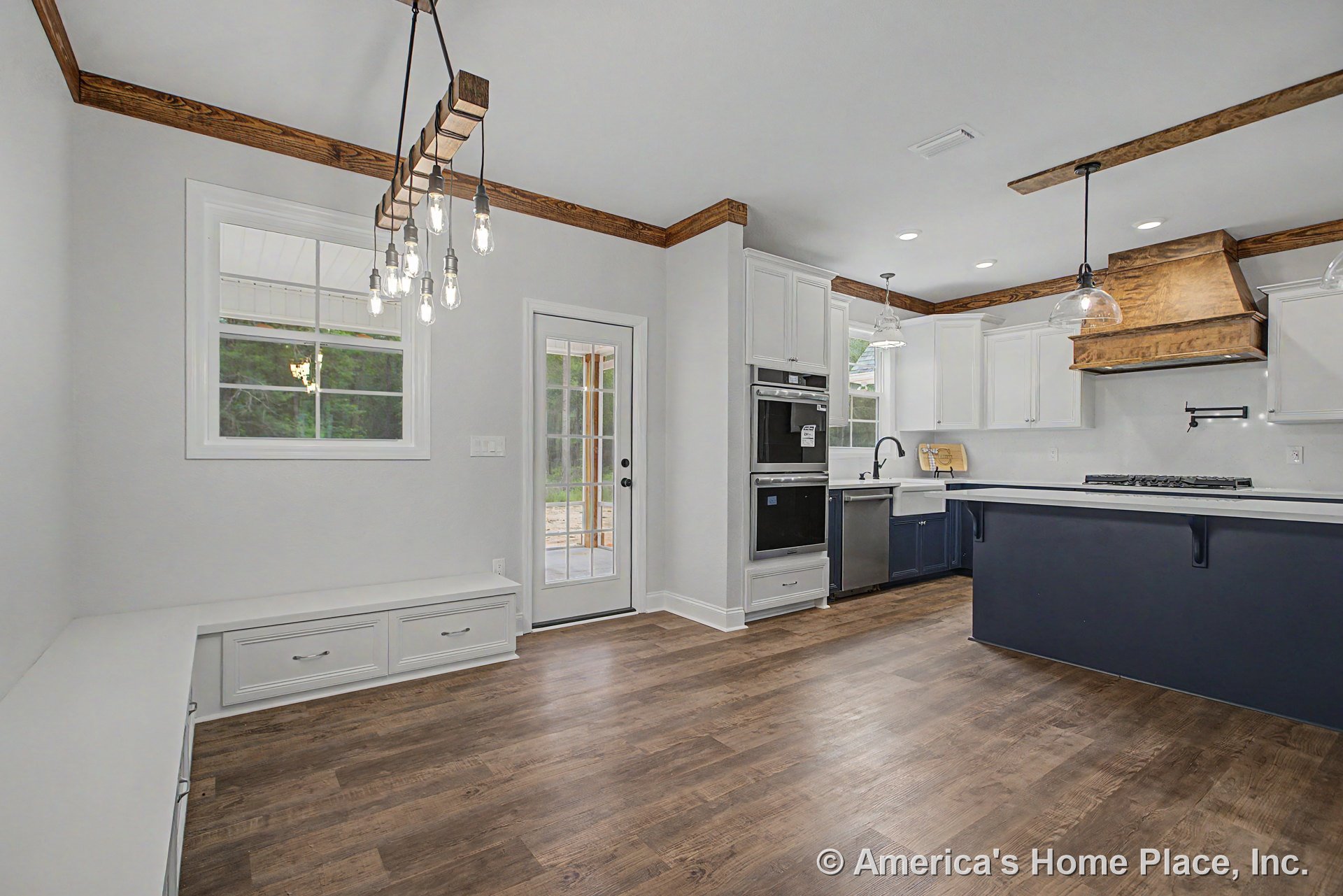 White kitchen with wood ceiling beams, custom wood range hood, double wall ovens, navy island topped with white quartz and pendant lights, farmhouse sink beneath window, built-in