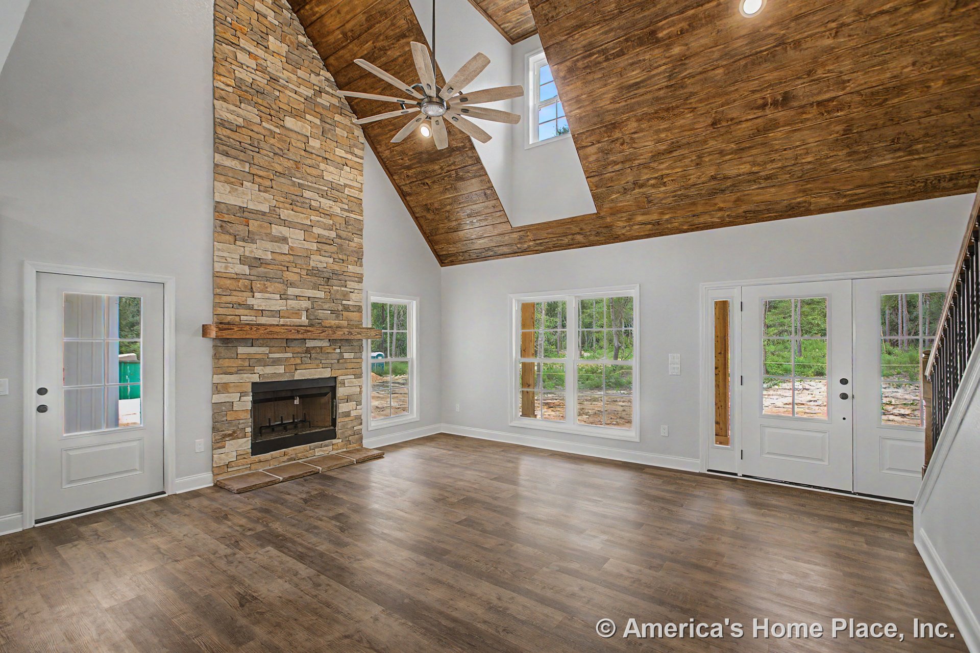 Vaulted wood ceiling with exposed beams, stone fireplace and mantel, wide plank wood flooring, expansive windows with white trim, double glass doors, ceiling fan featuring built-in