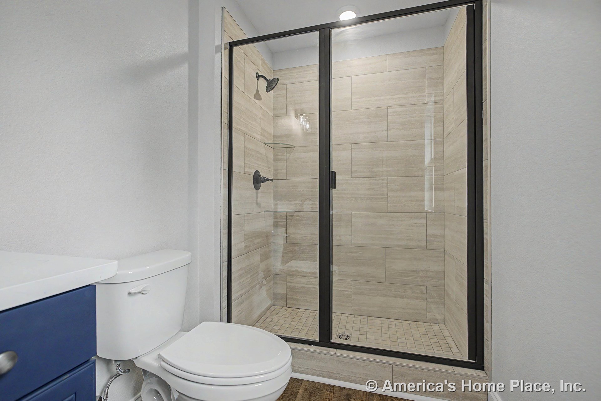 Walk-in shower with frameless glass door, large beige tile walls, built-in shower shelf, recessed ceiling light, white toilet, blue vanity cabinet with white countertop, modern