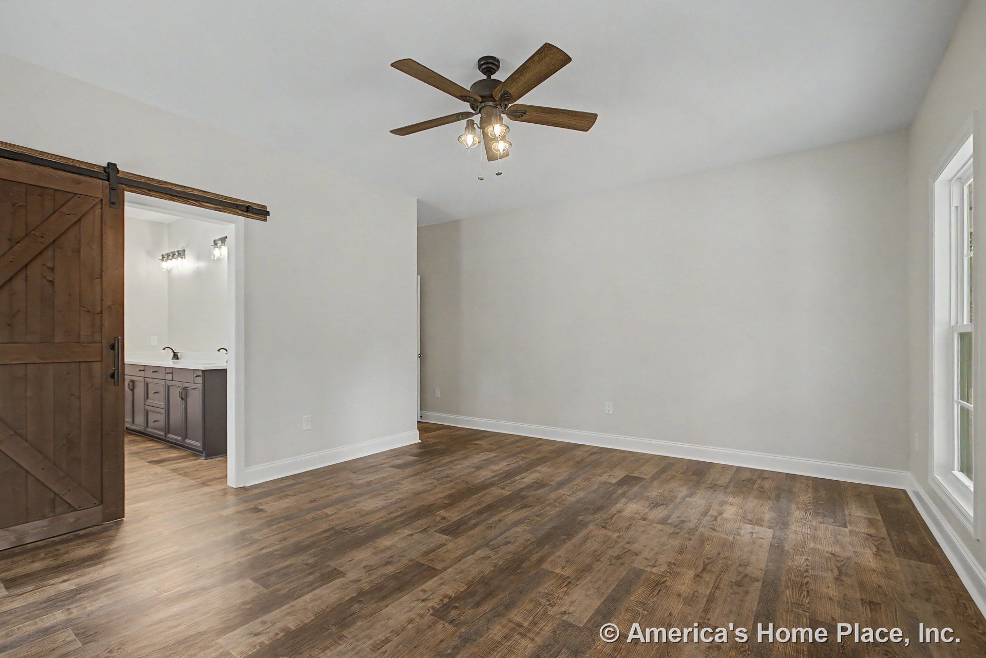Bedroom with wood-look plank flooring, ceiling fan with built-in lighting, large window, white baseboard trim, and sliding barn door opening to bathroom featuring double vanity and