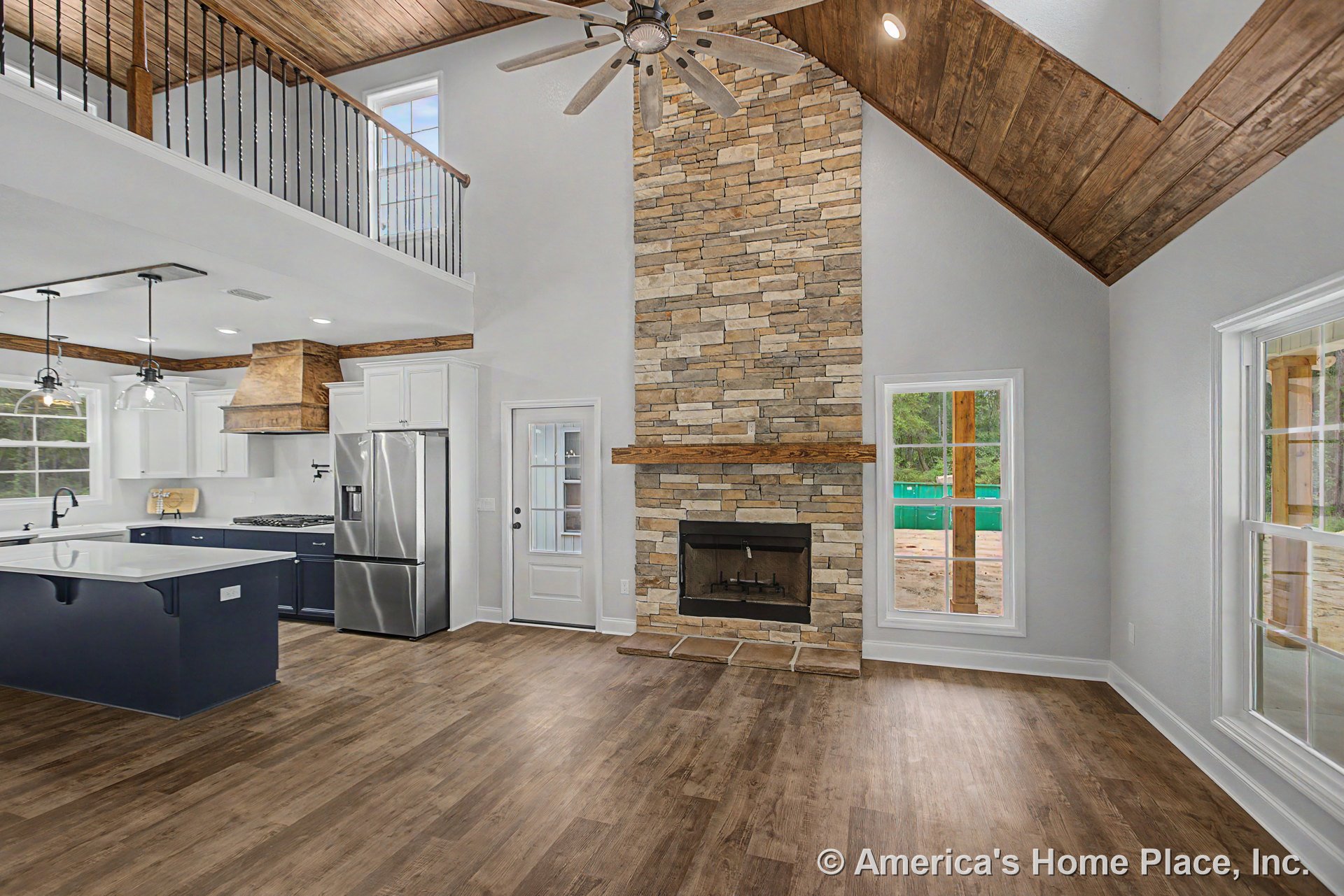 Tall stone fireplace with wood mantel anchors open concept living area, featuring vaulted wood plank ceiling, large windows with white trim, wood flooring, and adjacent kitchen