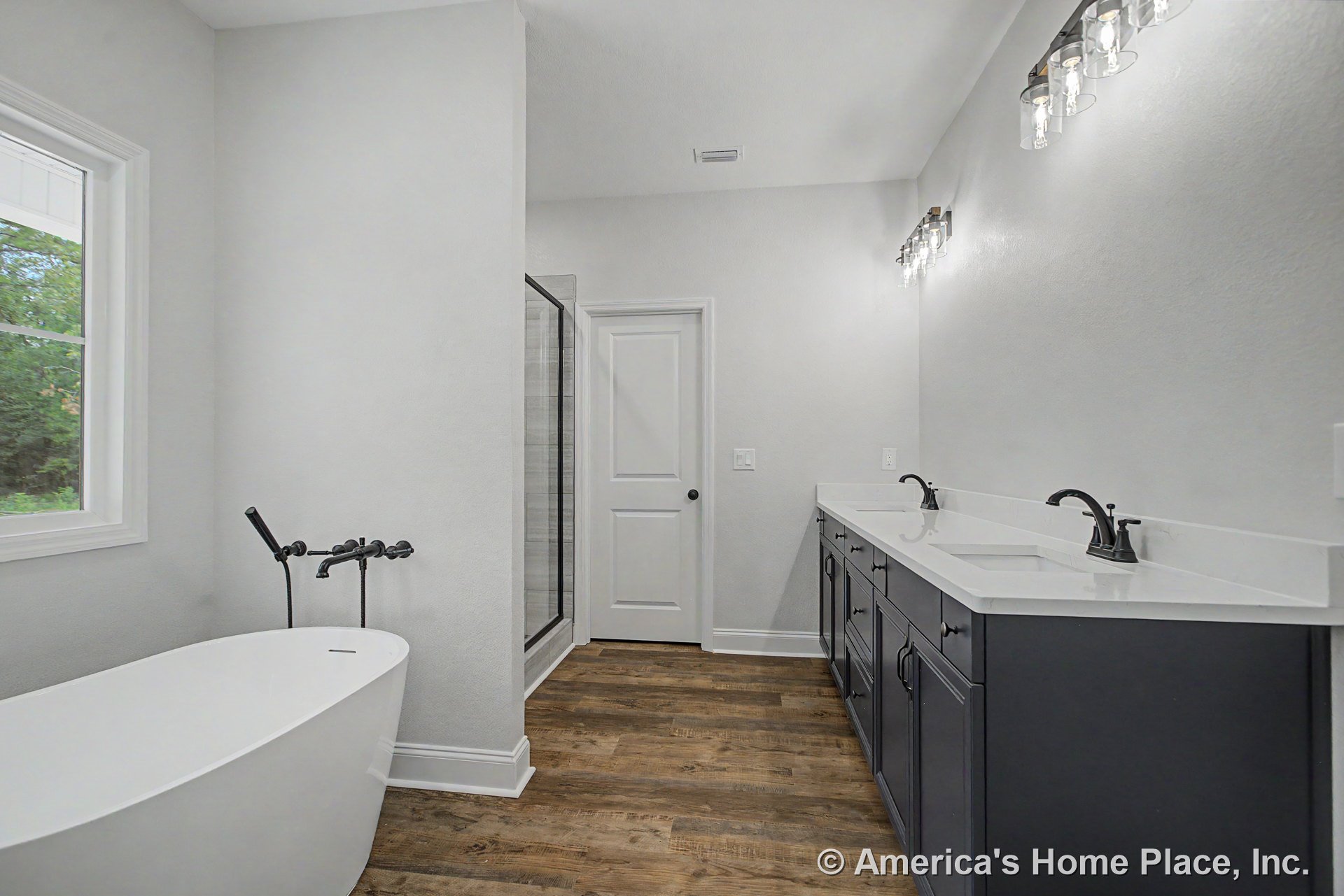 Freestanding soaking tub beside double sink vanity with quartz countertops, wood-look plank flooring, frameless glass shower enclosure, matte black fixtures, modern wall-mounted