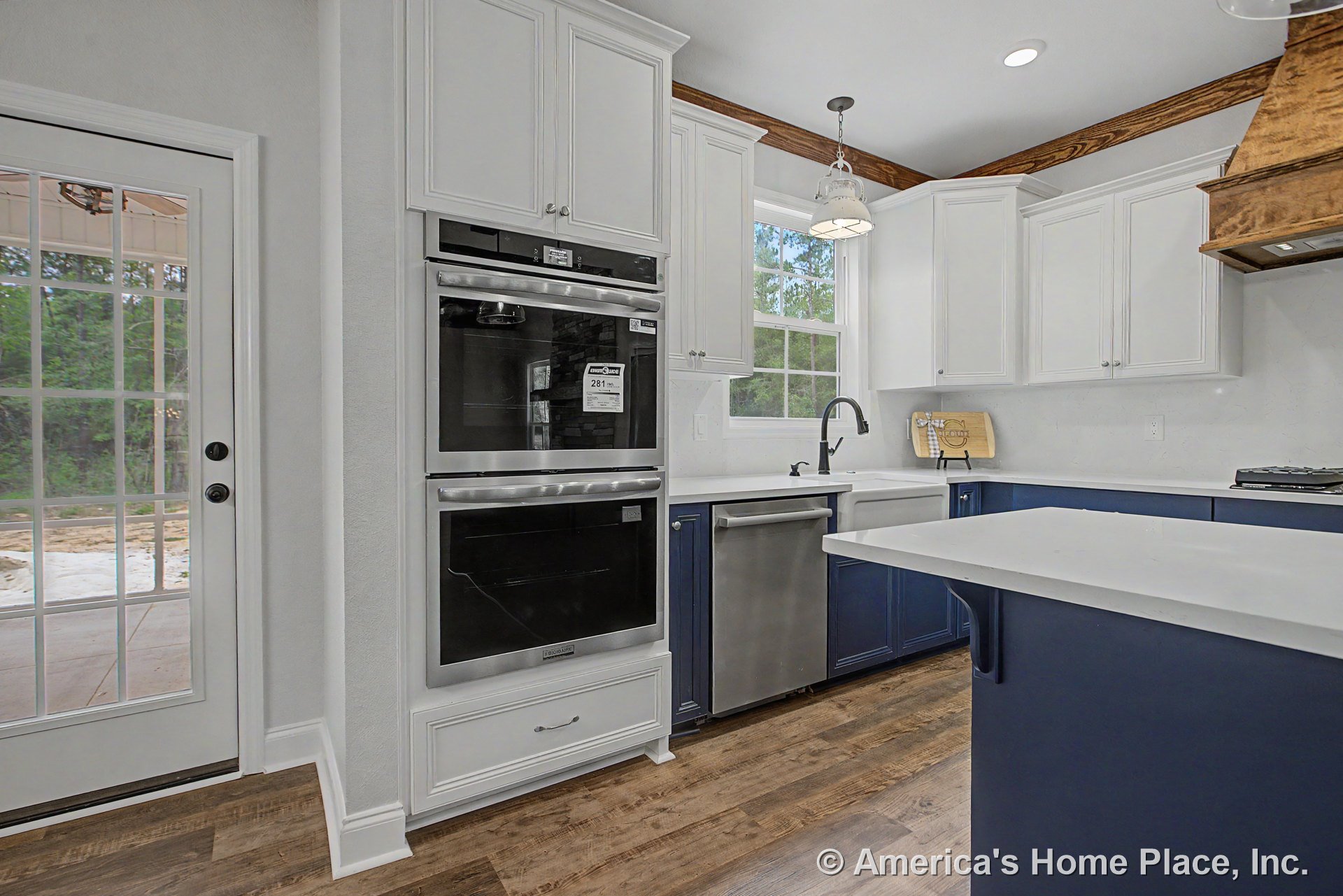 White shaker upper cabinets and blue base cabinetry with quartz countertops, double wall ovens, farmhouse sink, stainless dishwasher, wood-look flooring, pendant lighting, window