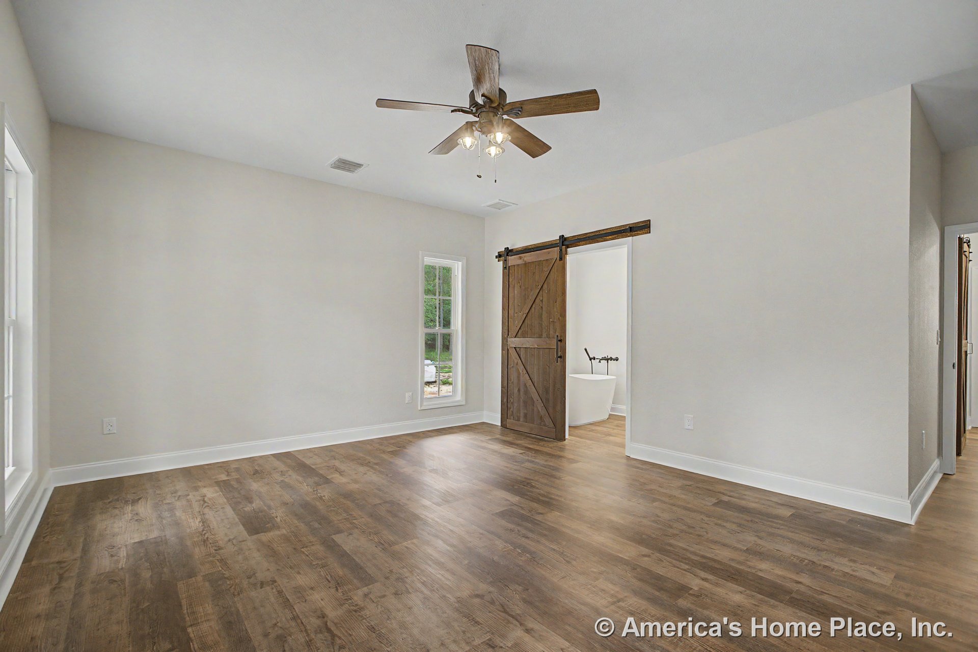 Bedroom with wood-look plank flooring, ceiling fan and light fixture, white trim and baseboards, sliding barn door to bathroom, large window, and bathroom entry featuring a