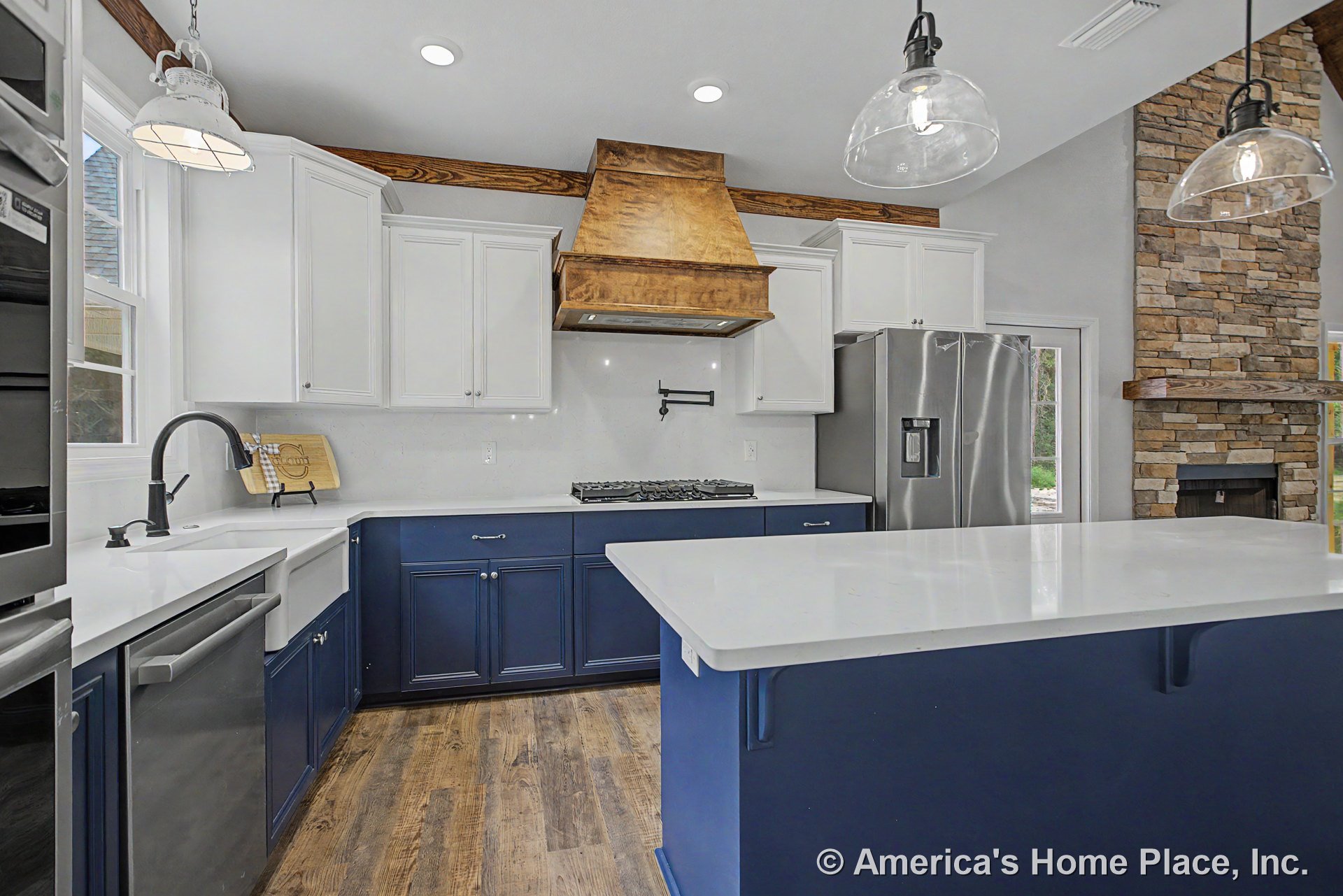 Blue lower cabinets and white upper cabinets with quartz countertops, wood range hood above built-in oven, farmhouse sink, stainless steel appliances, pot filler above stove
