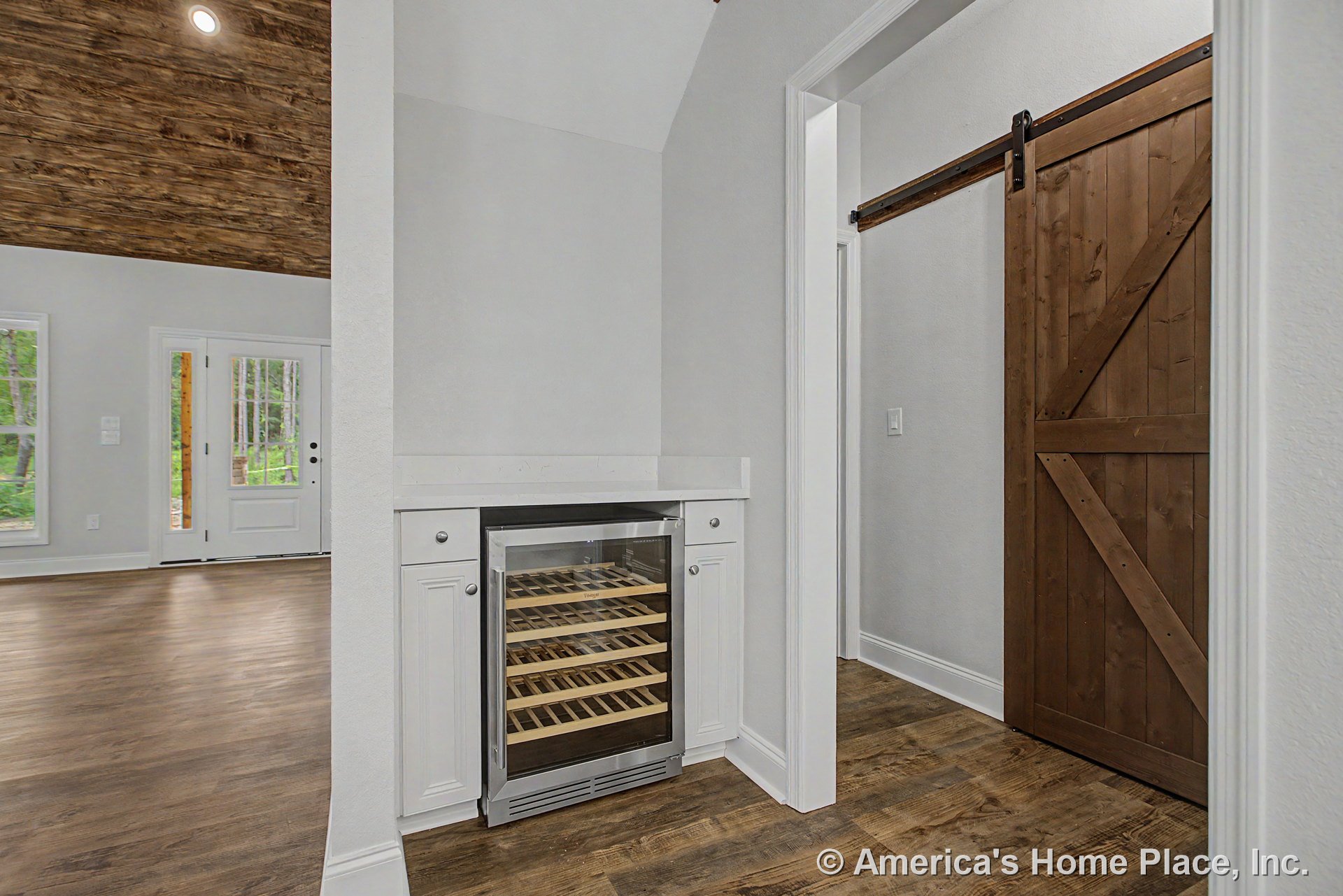 Built-in beverage cooler with glass door integrated into white shaker cabinetry, quartz countertop, adjacent wood sliding barn door, wood-look flooring, vaulted wood plank ceiling