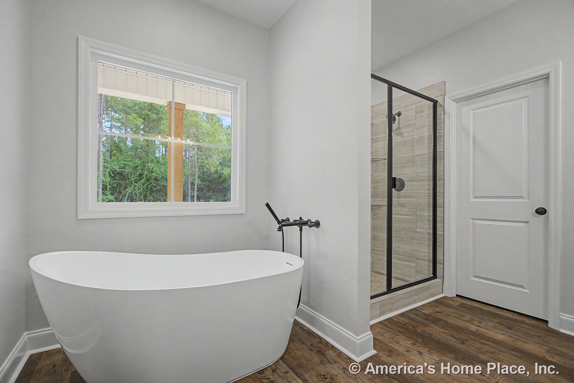 Freestanding soaking tub with matte black faucet beside a large window with trim, glass-enclosed shower with tile walls, wood-look plank flooring, white paneled door, baseboard