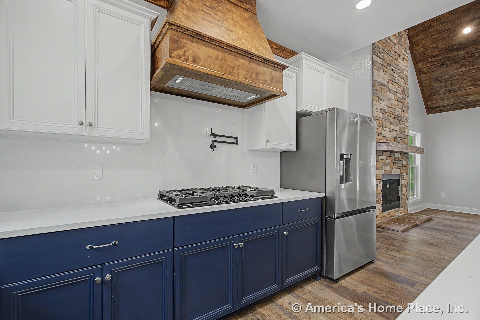 Custom kitchen with wood vent hood, white upper and blue lower cabinetry, quartz countertops, gas cooktop with pot filler, stainless refrigerator, vaulted ceiling, hardwood