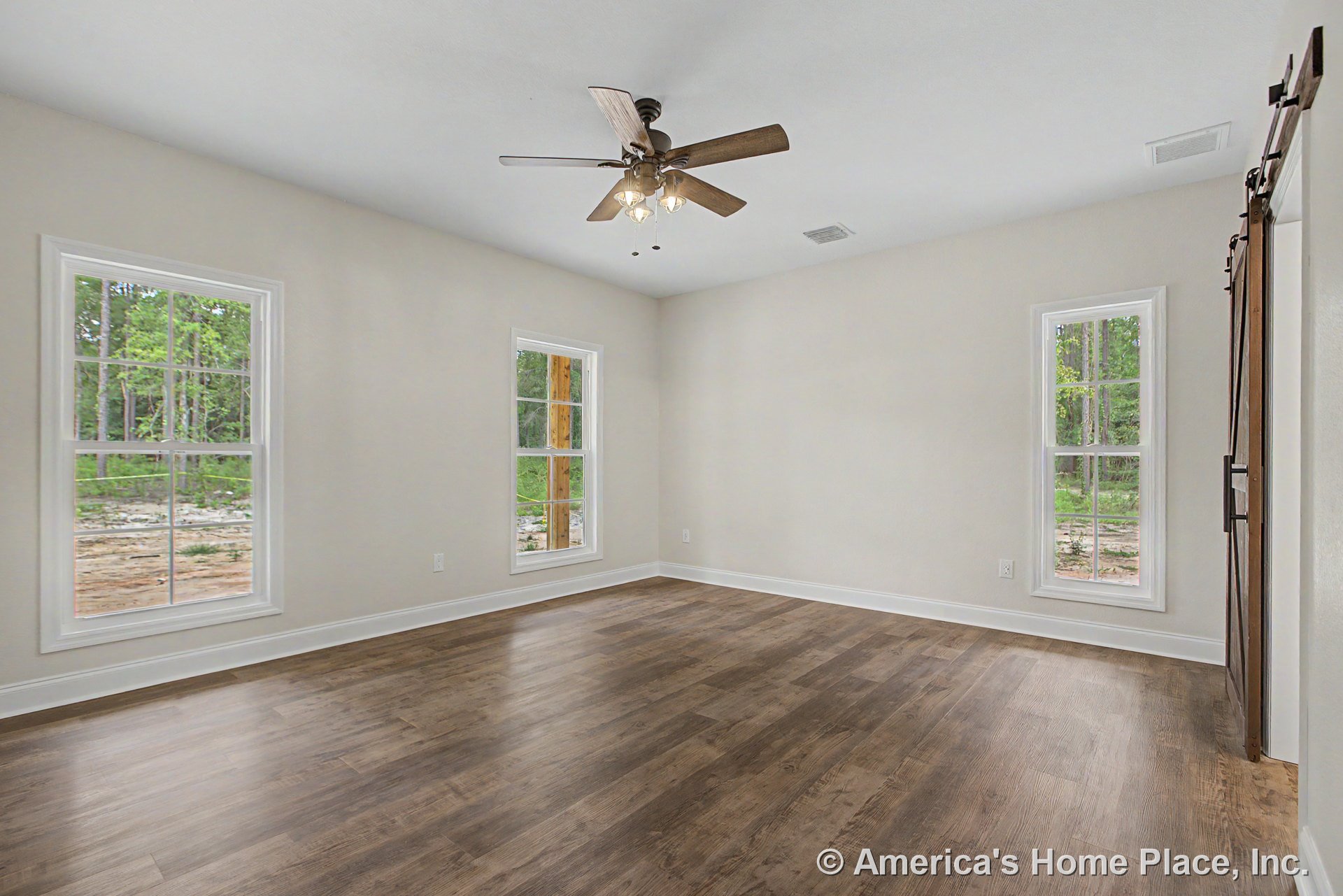 Wood-look plank flooring, three tall windows with abundant natural light, ceiling fan with integrated light fixture, sliding barn door, white baseboard trim, and neutral wall color