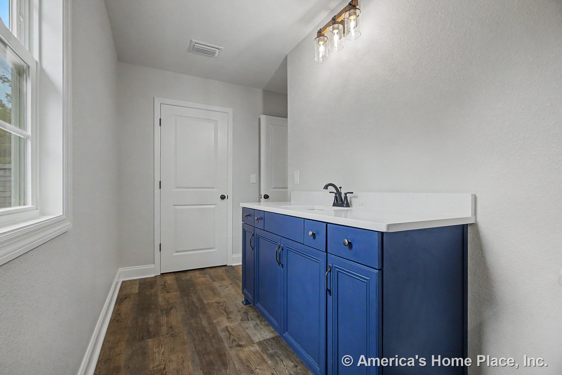 Blue bathroom vanity with white solid surface countertop, three-light wall fixture above, wood-look vinyl flooring, white trim and doors, large window with casing.