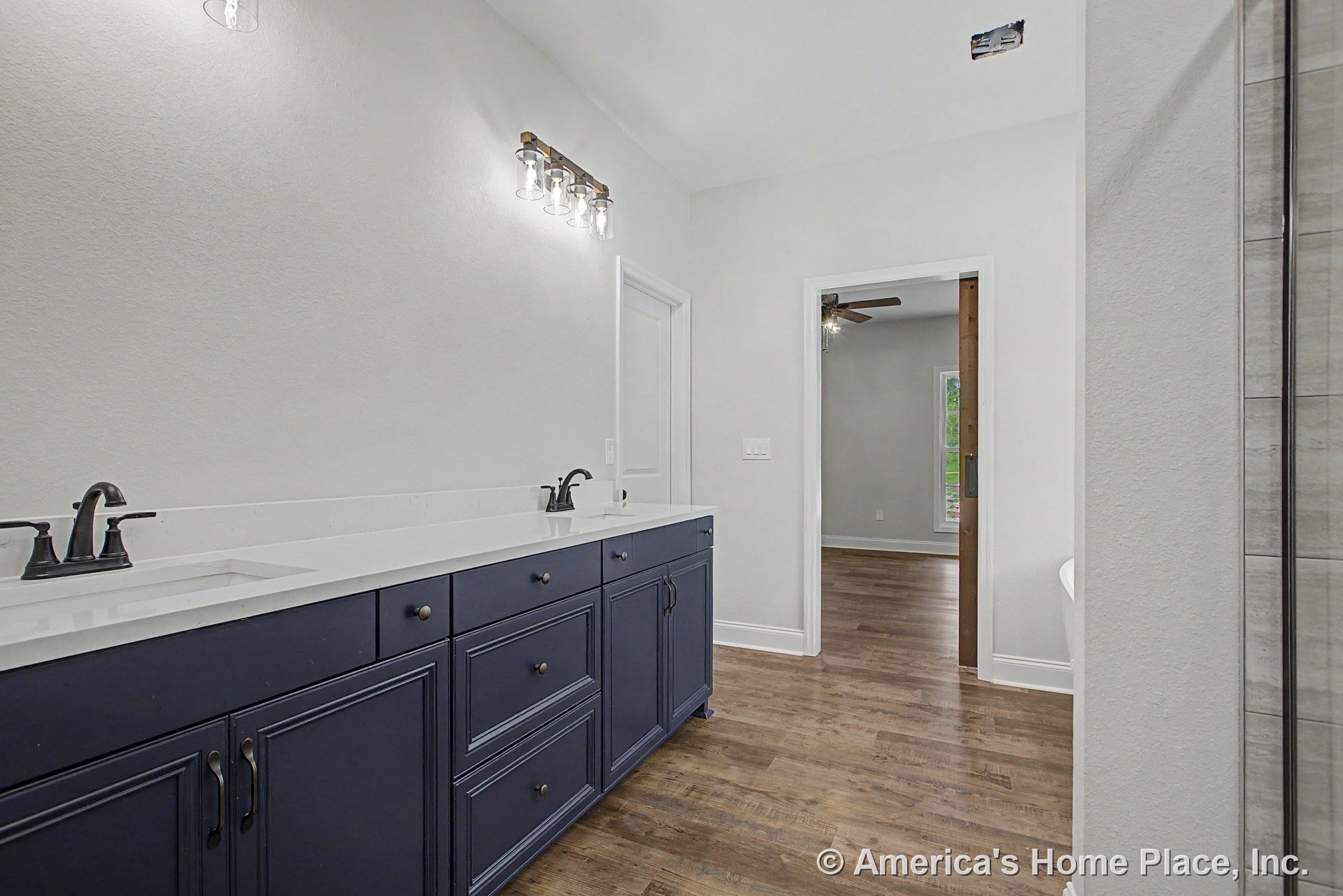 Double vanity with navy cabinetry and white quartz countertops, matte black faucets, wall-mounted sconce lighting, wood-look flooring, undermount sinks, white walls, and doorway