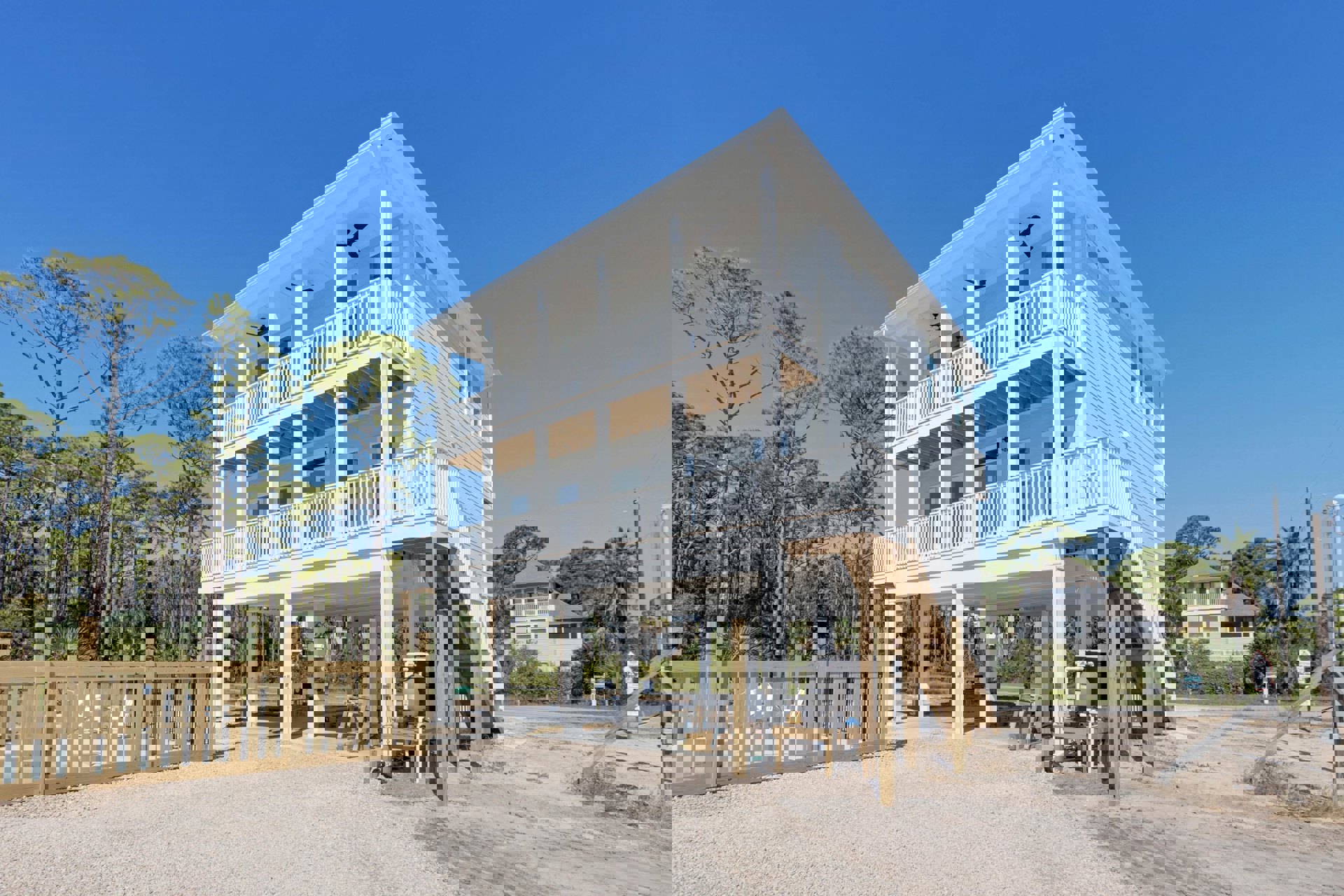 White two-story house with balcony, wood deck and stairs, wooden fence enclosing porch area, ceiling fan with light fixture, outdoor chairs, trees and sky in background