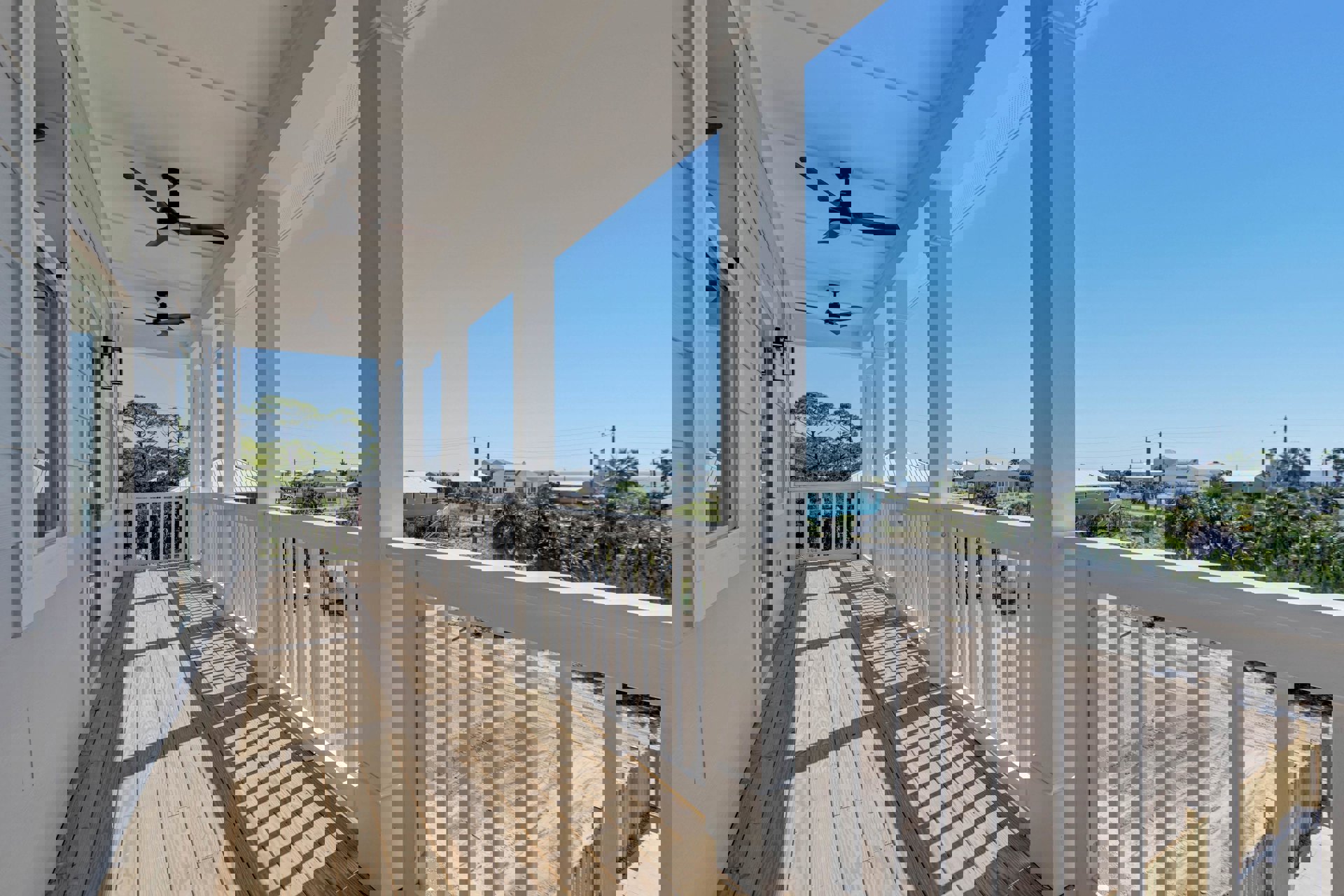 Spacious porch with wooden deck, white railings and pillars, ceiling fan with light, overlooking neighboring houses and sky