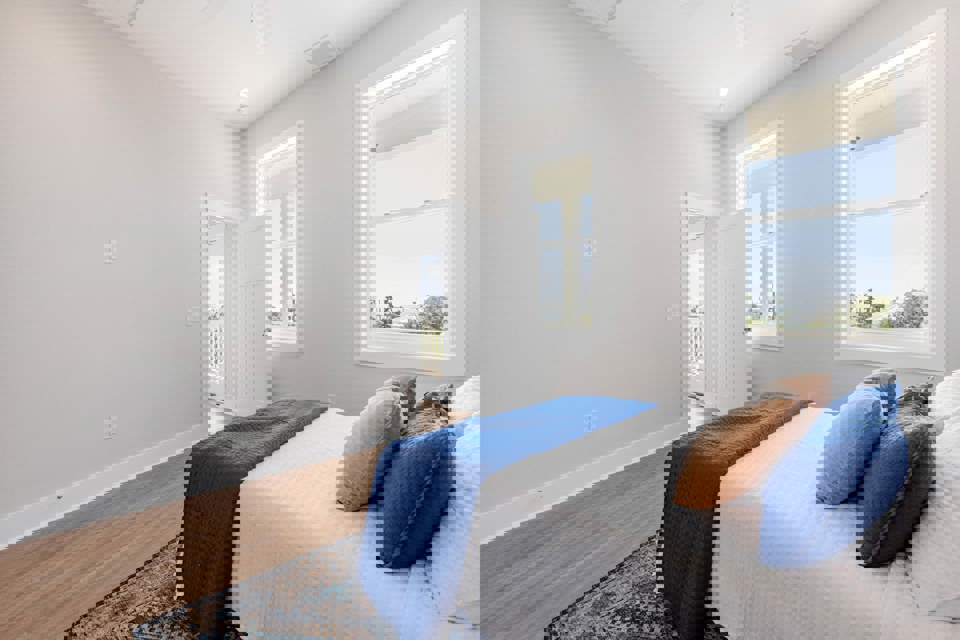 Bedroom with blue bedspread, assorted pillows in blue and brown, patterned rug, white walls, window showing neighboring house, and door to the side