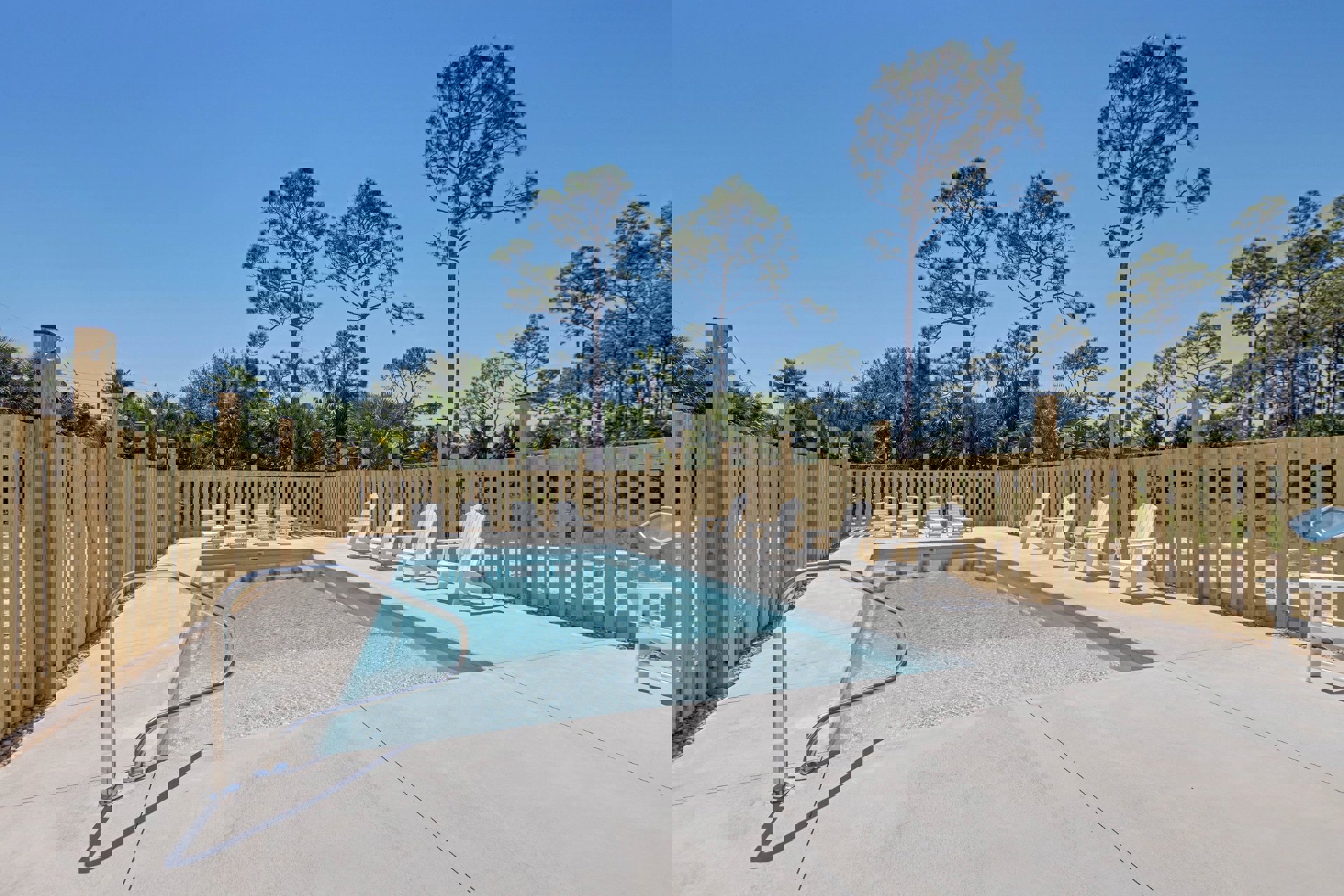 Rectangular swimming pool with metal handrail, white patio chairs beside a wooden fence, group of trees and clear blue sky in the background