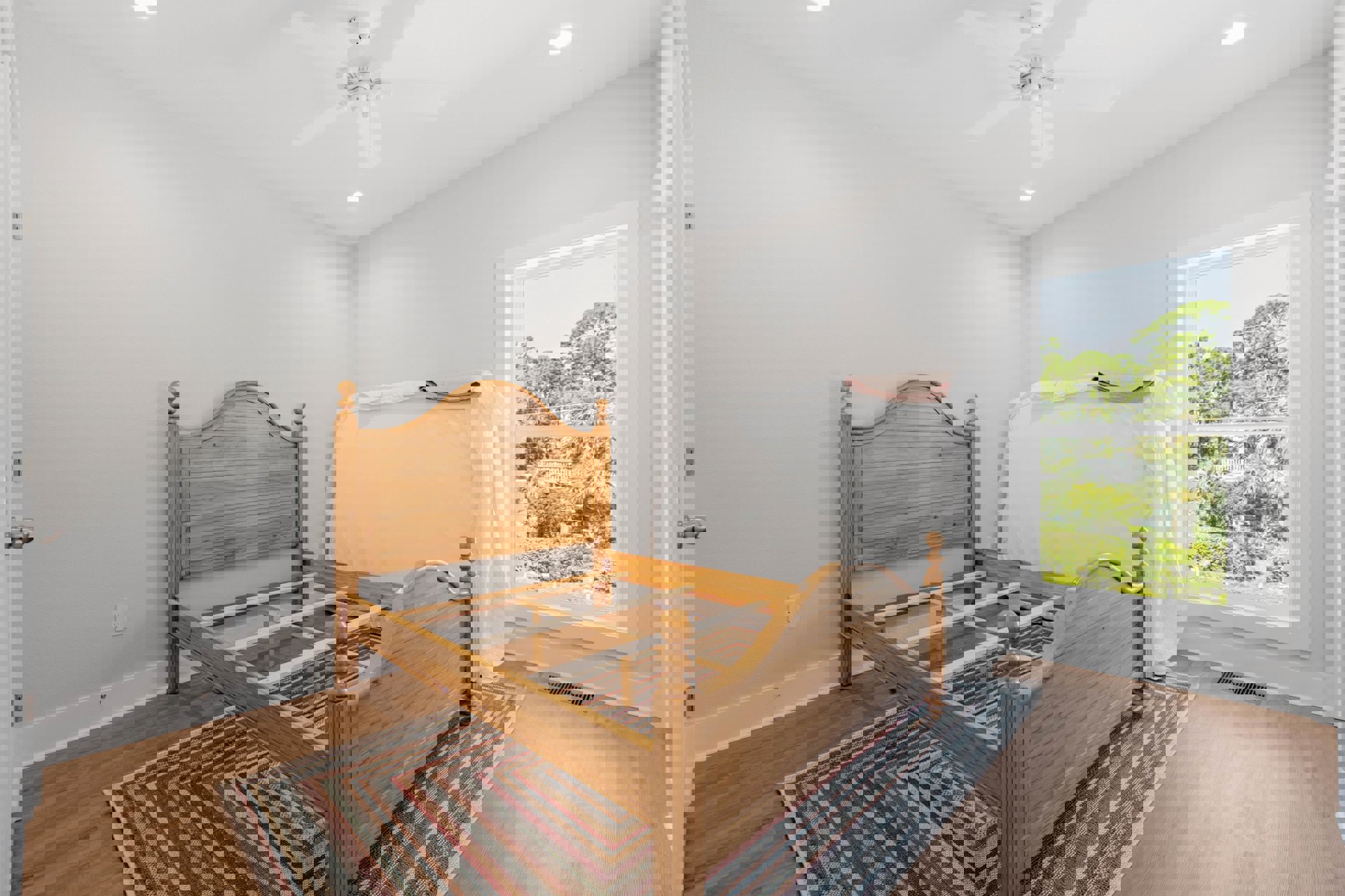 Wooden bed frame with white sheet and headboard against wall, large window showing trees outside, rug covering hardwood floor in bedroom.