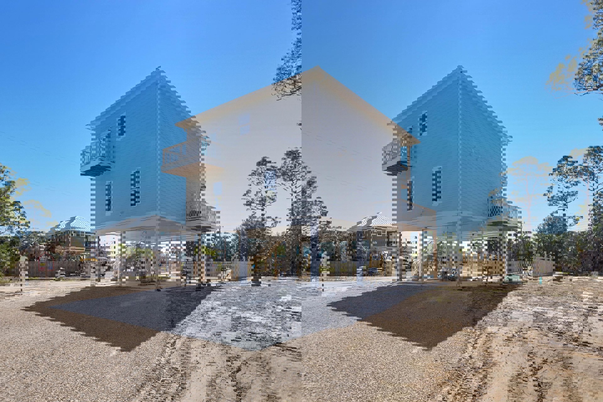 Two-story house under construction with exposed framing, gravel driveway, tire tracks in dirt, and mature trees surrounding the property
