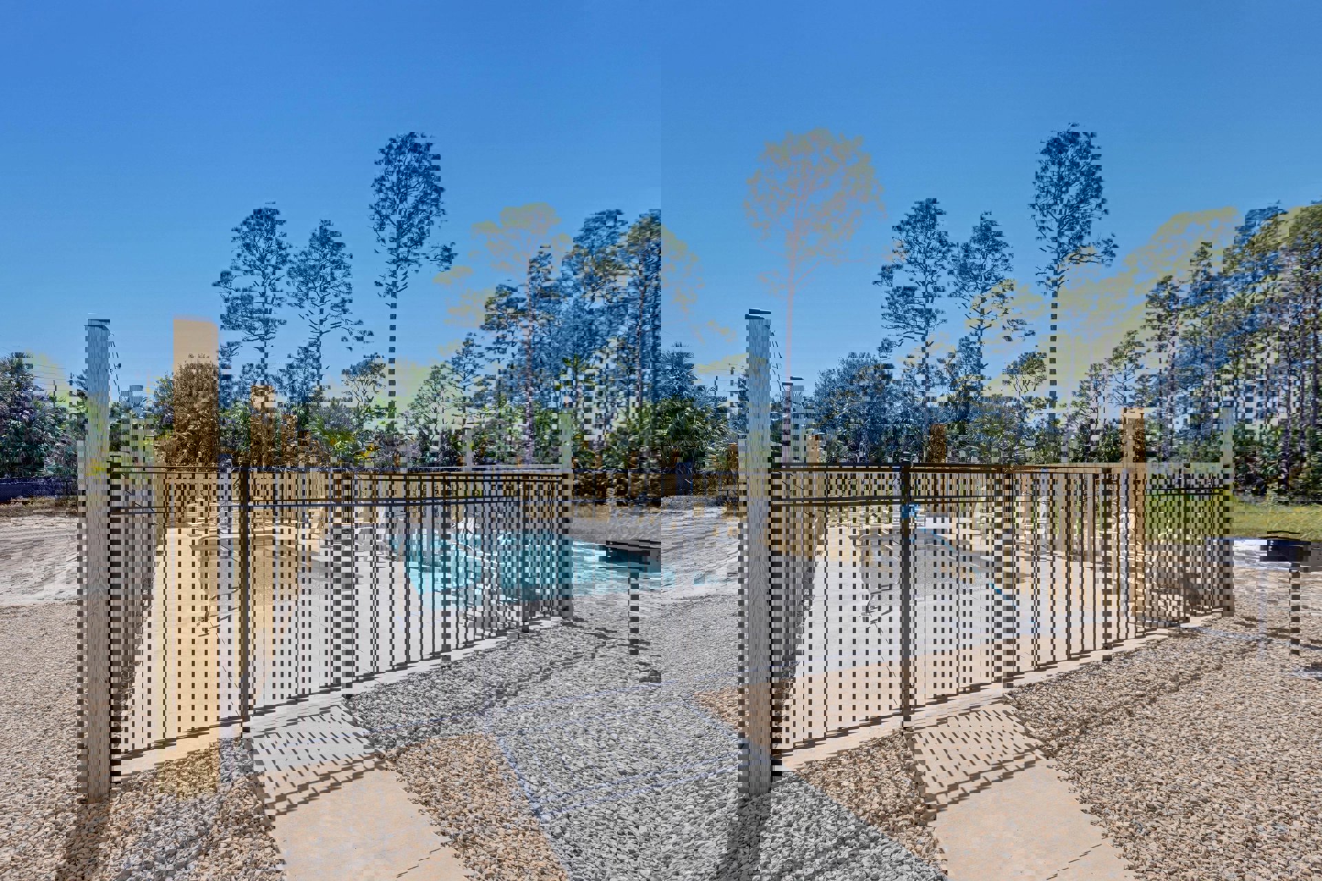Backyard pool surrounded by metal fence, concrete walkway, and landscaping rocks, with tall trees and blue sky in the background
