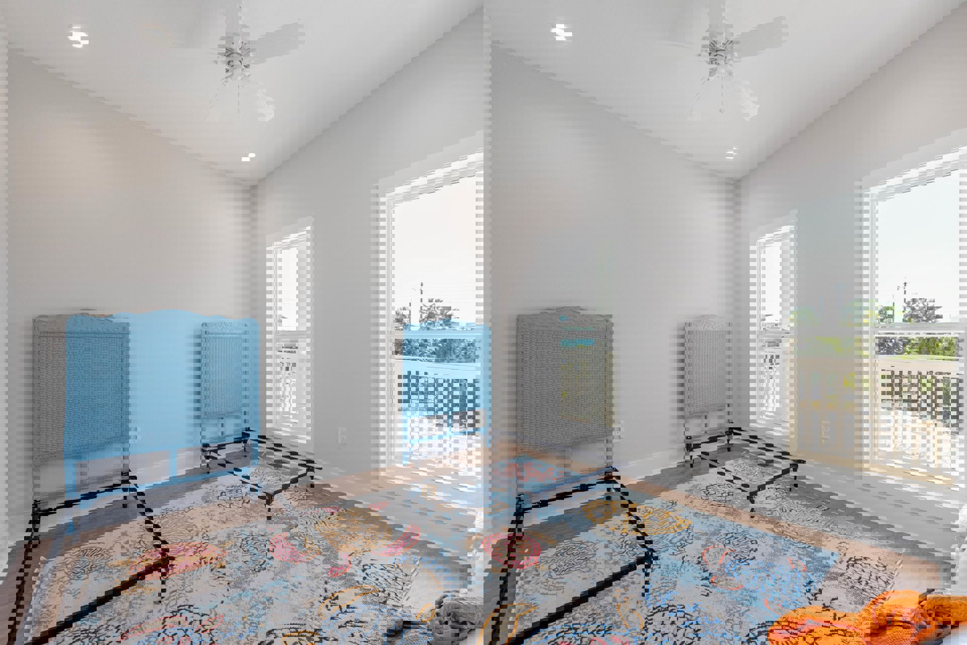 Bedroom with blue woven bed frame and rattan headboard, white walls, blue chairs, white ceiling fan, window with white railing overlooking palm trees, patterned blanket, and