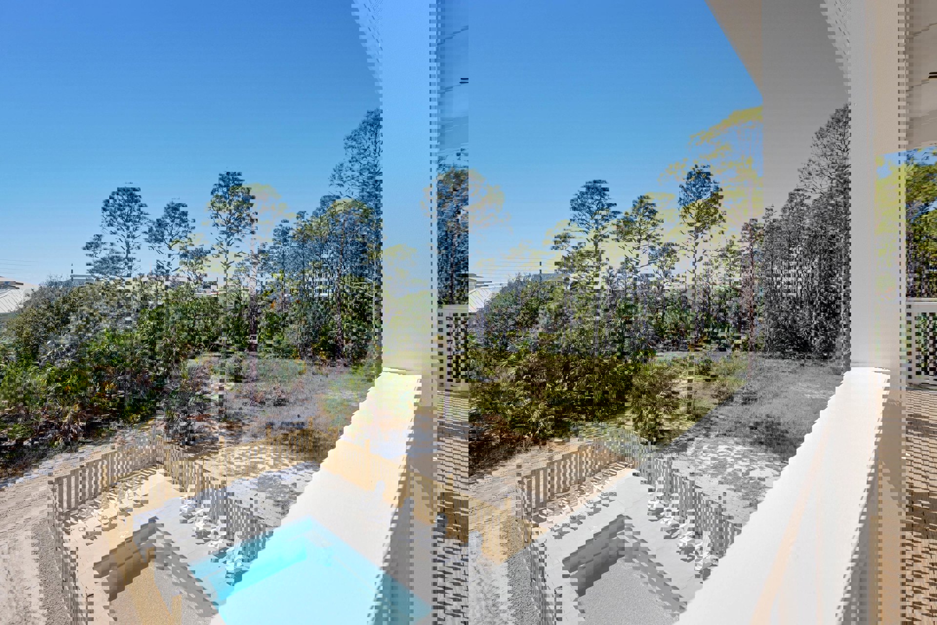 Concrete poolside with white chairs, tall trees, and metal fence surrounding a residential backyard pool; modern exterior building visible in background.