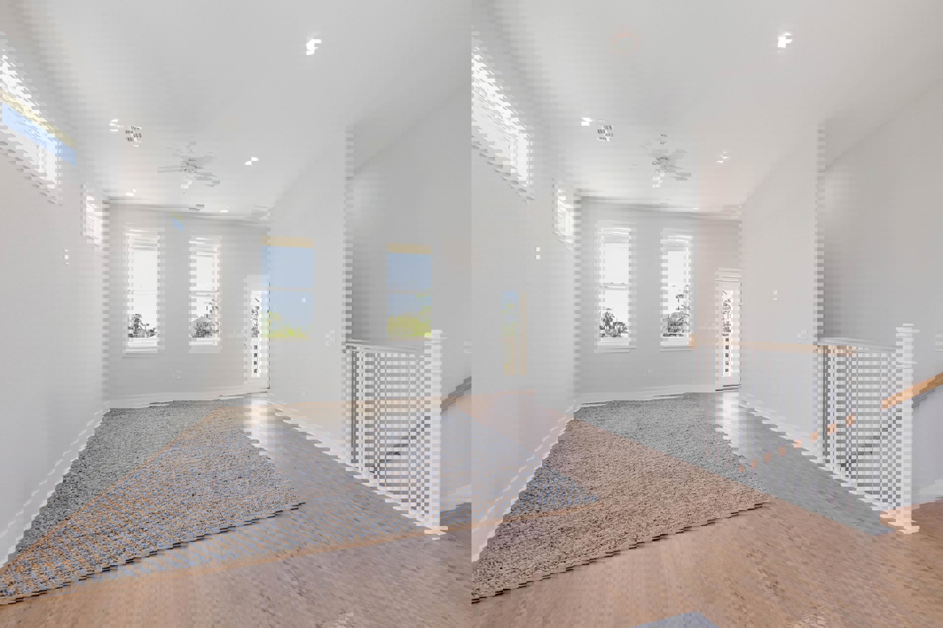 Living room with wide plank wood flooring, patterned area rug, large window overlooking trees and neighboring house, ceiling fan, neutral plaster walls.