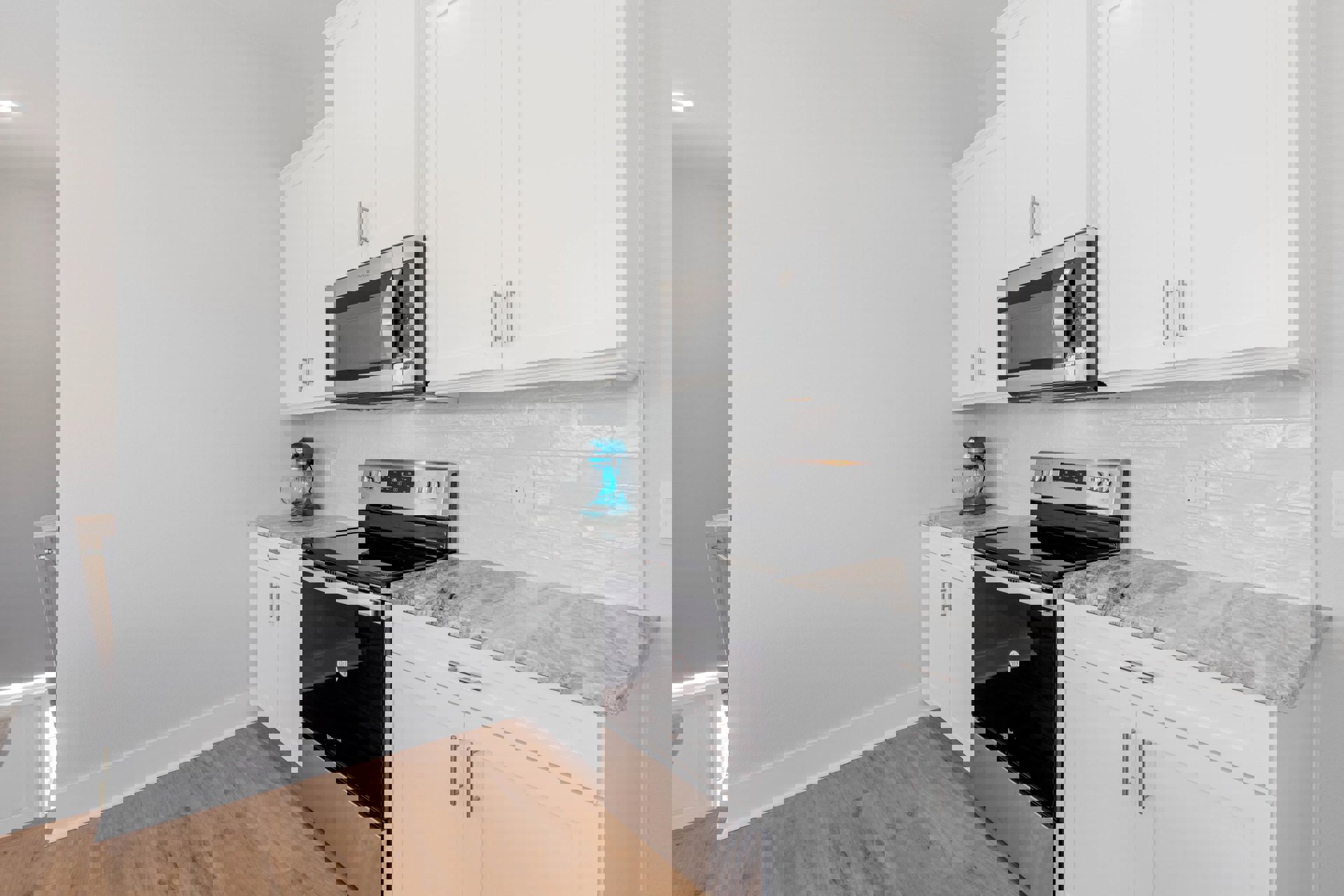 White kitchen cabinets, stainless steel stove and oven, built-in microwave, blue stand mixer with glass bowl on white countertop, wood flooring with white baseboard, white walls.