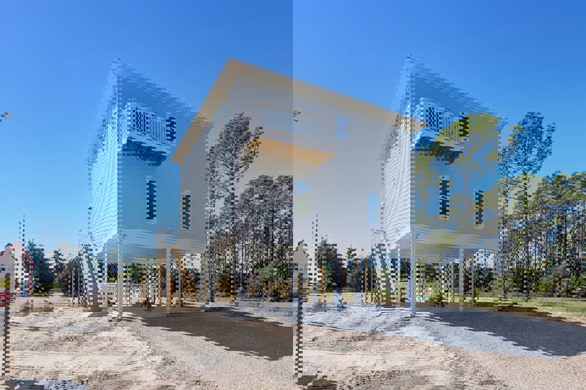 Two-story wood-framed house under construction with exposed plywood, white-framed windows, elevated deck and stairs, surrounded by tall trees