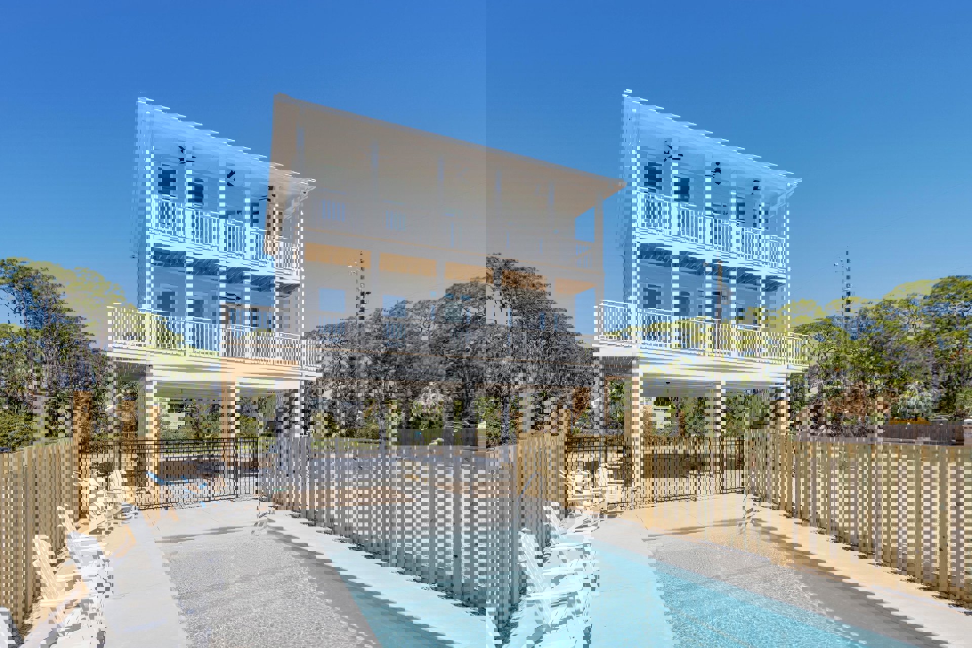 White two-story house with balcony, fenced backyard, rectangular swimming pool, row of white patio chairs, and surrounding trees under blue sky