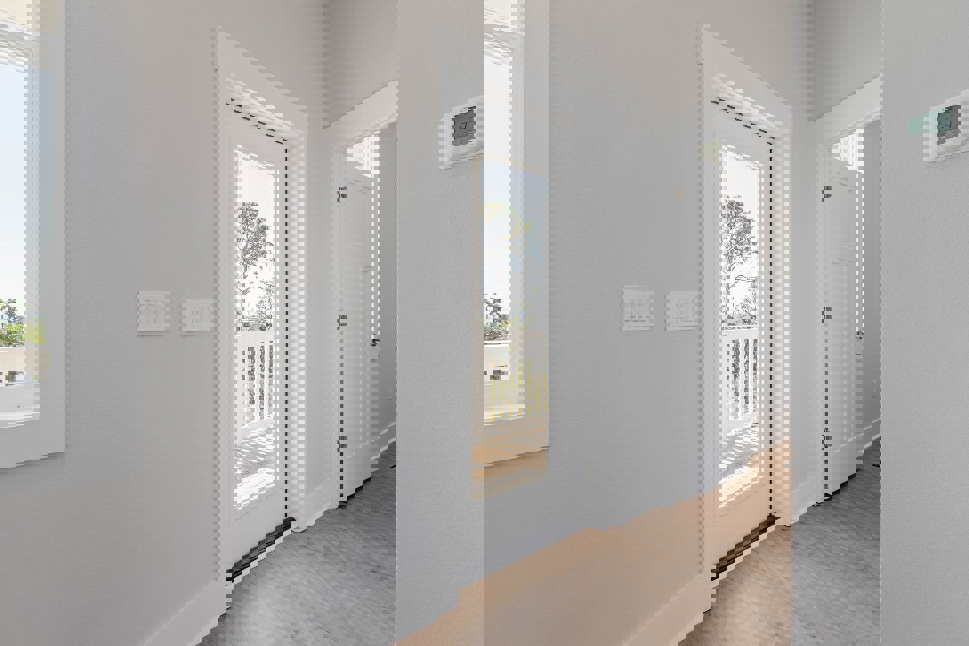White door with glass panel, wood laminate flooring, white wall with row of light switches, close-up of thermostat, white railing, leafy trees visible outside