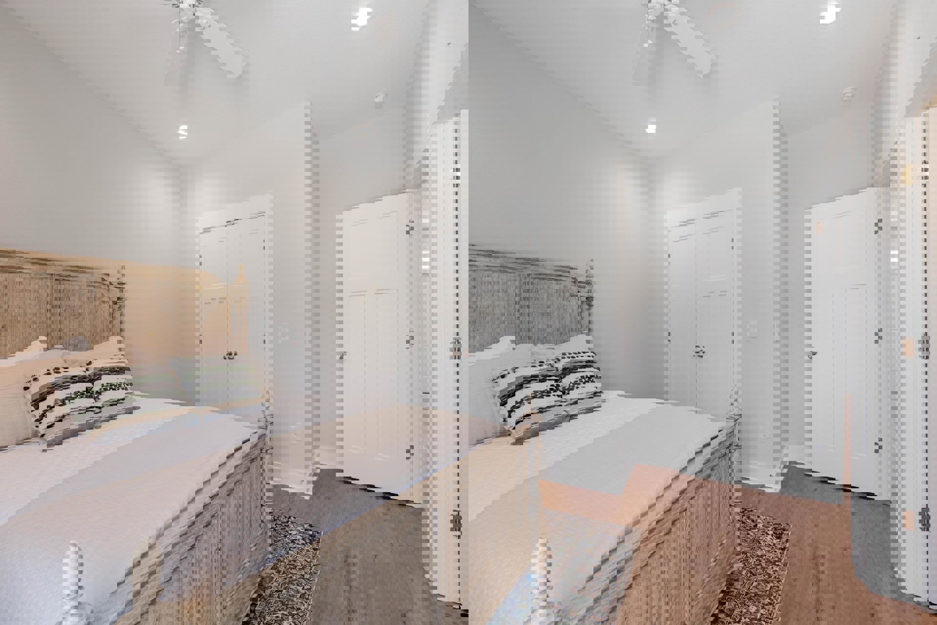 Bedroom with a wooden bed frame, white bedding and pillows, ceiling fan above, white door with silver knob, neutral walls, and stone decor accent