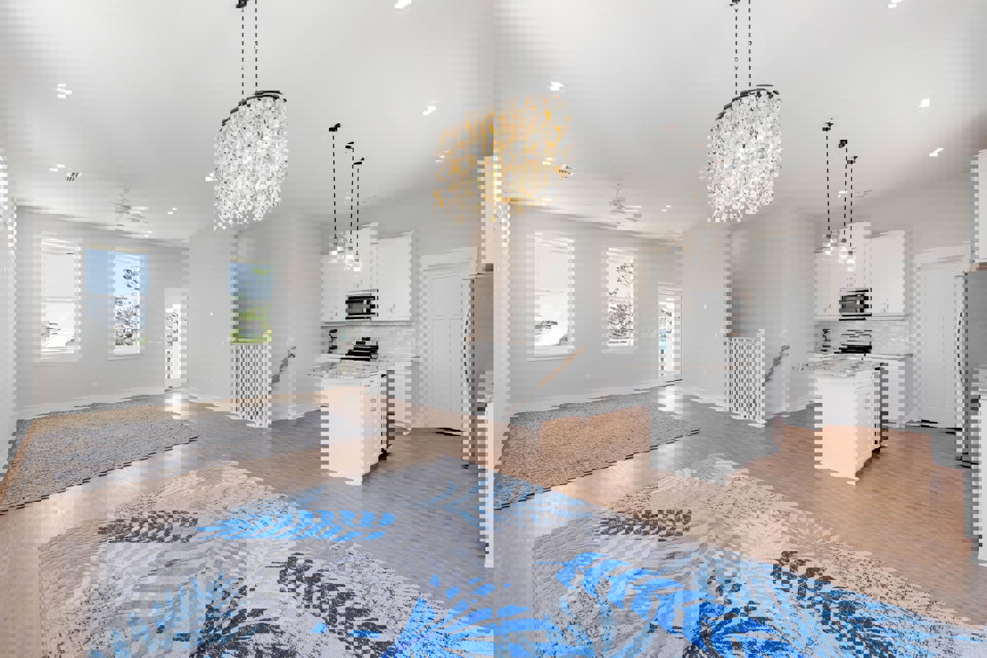 Spacious living room featuring oyster shell chandelier, patterned rug on hardwood floor, large windows with tree and rooftop views, white cabinetry, and built-in microwave oven