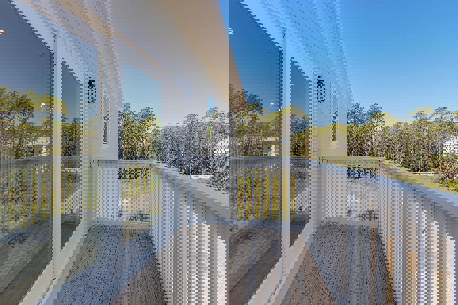 Wooden deck with white railing, glass door, and adjacent window; outdoor light fixture in glass case; trees and sky visible in background