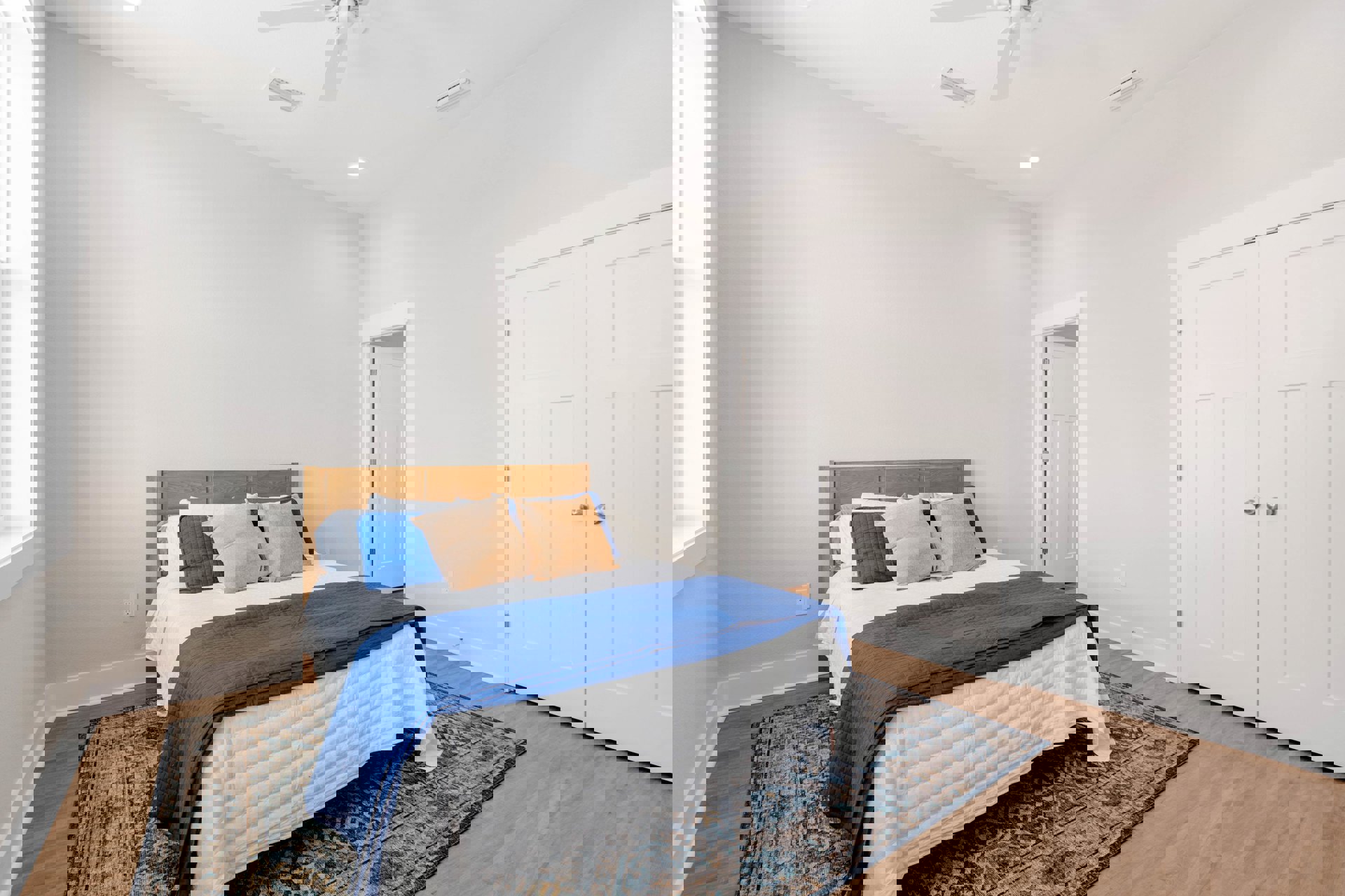 Queen bed with blue and white blankets, white pillows, light wood floor, white walls, and a white door with silver knobs in a modern bedroom.