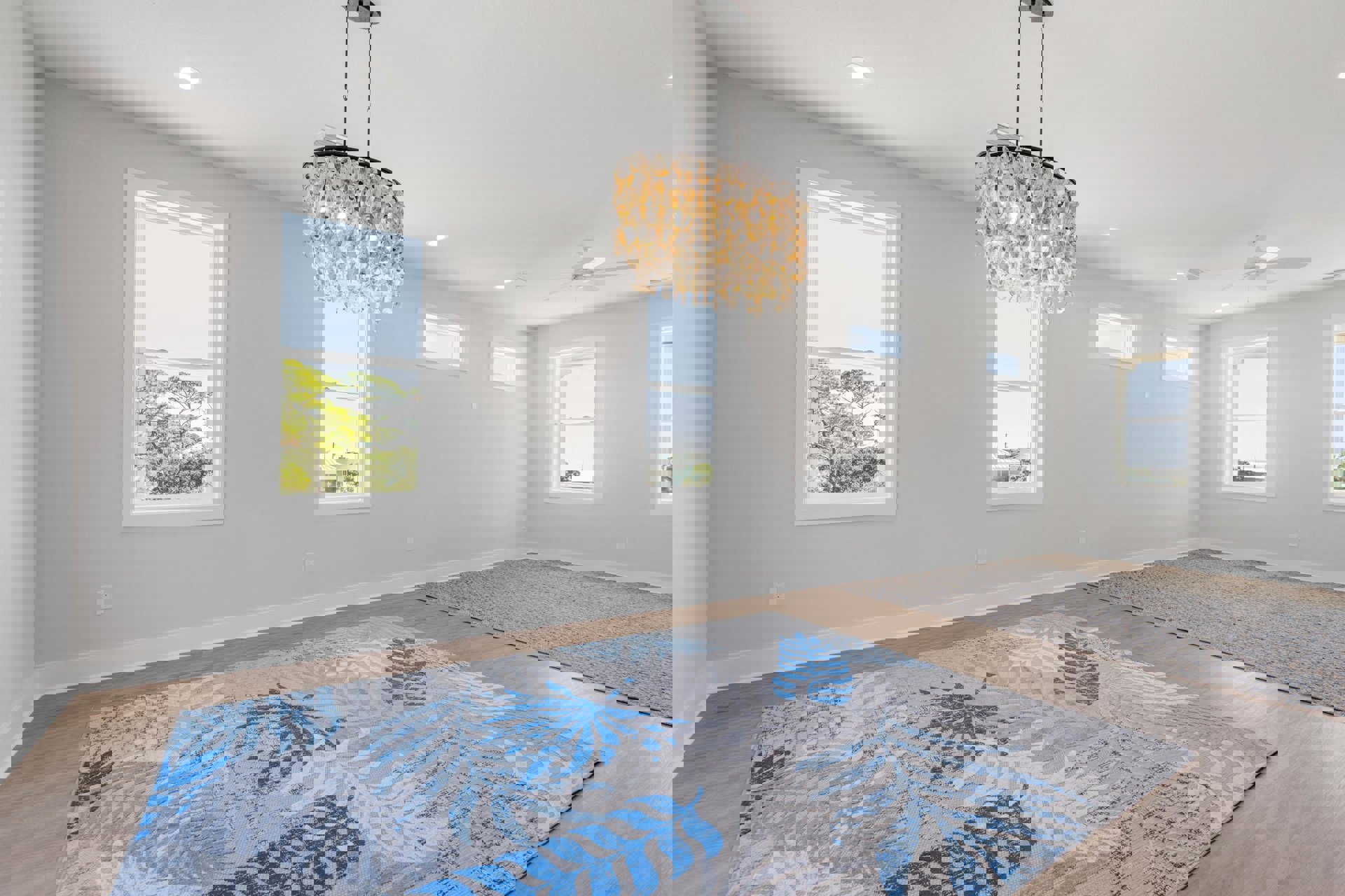 Living room with oyster shell chandelier, patterned rug on hardwood floor, large windows showing trees and cityscape, white walls, crown molding, and ceiling fixture.