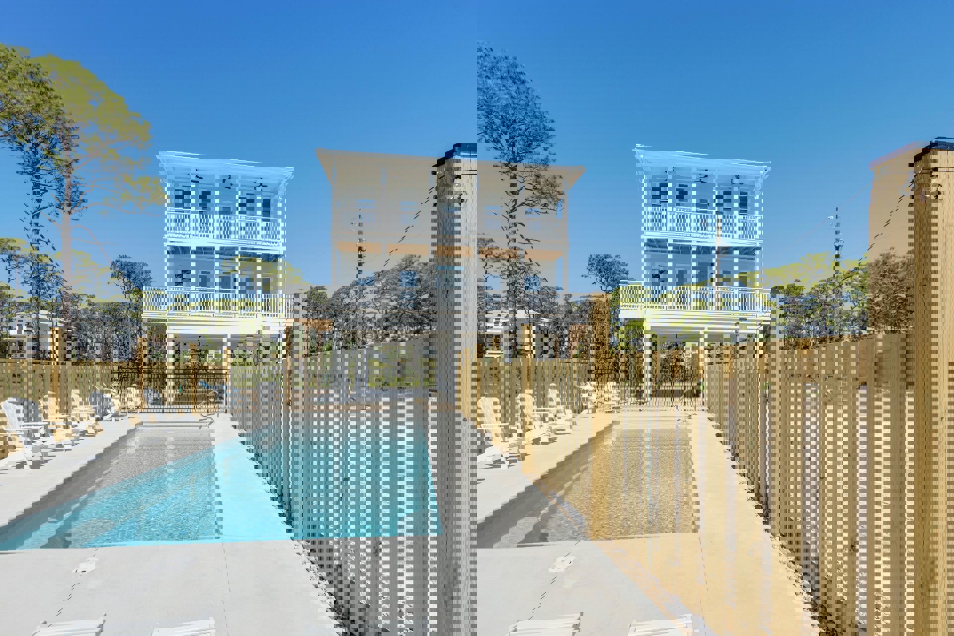 Rectangular swimming pool bordered by white lounge chairs, wooden privacy fence, and modern white house with large windows and attached porch.