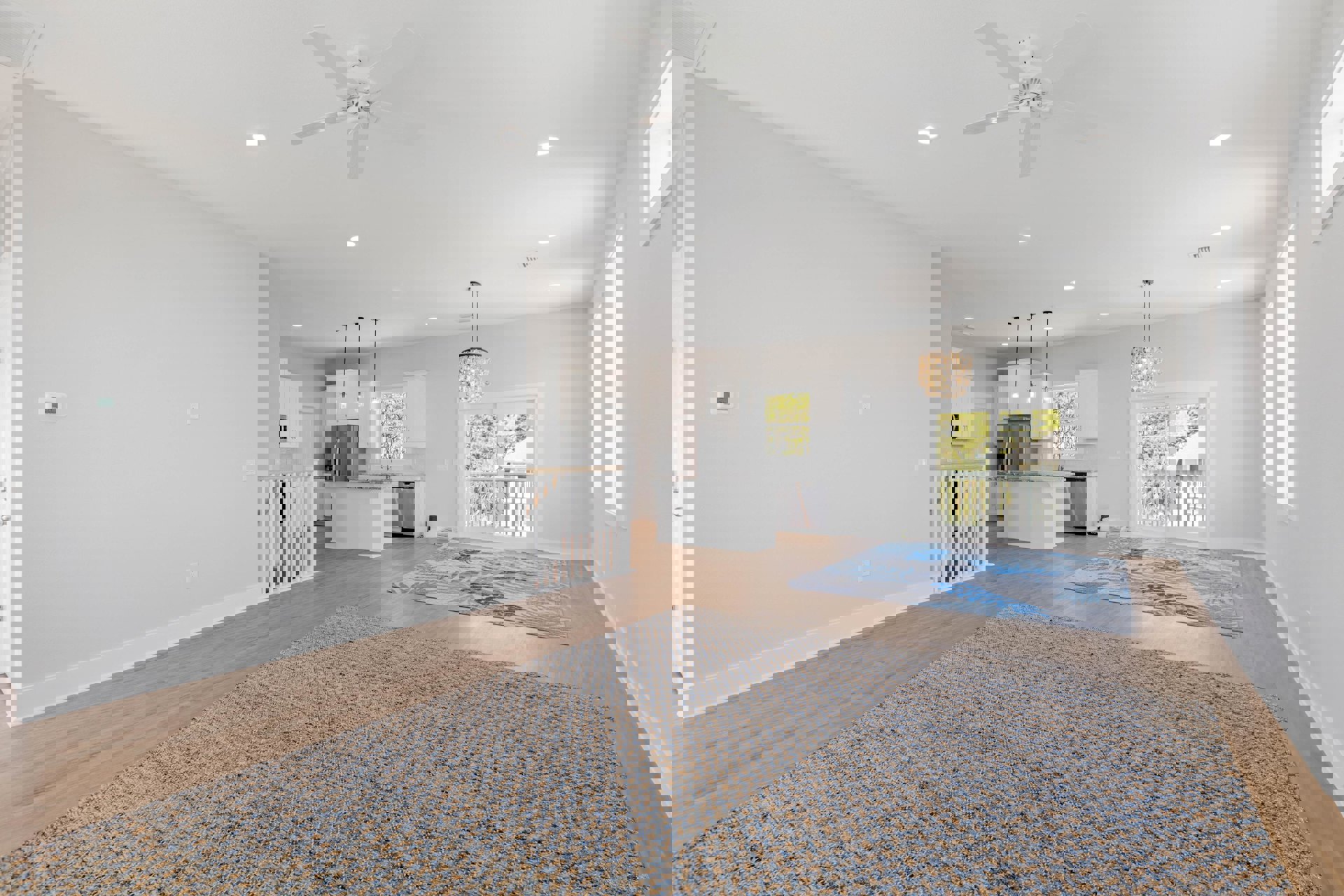 Open living area with laminate wood flooring, patterned rug, ceiling fan, white plaster walls, staircase with wood railing, and sliding glass door leading to outdoor space