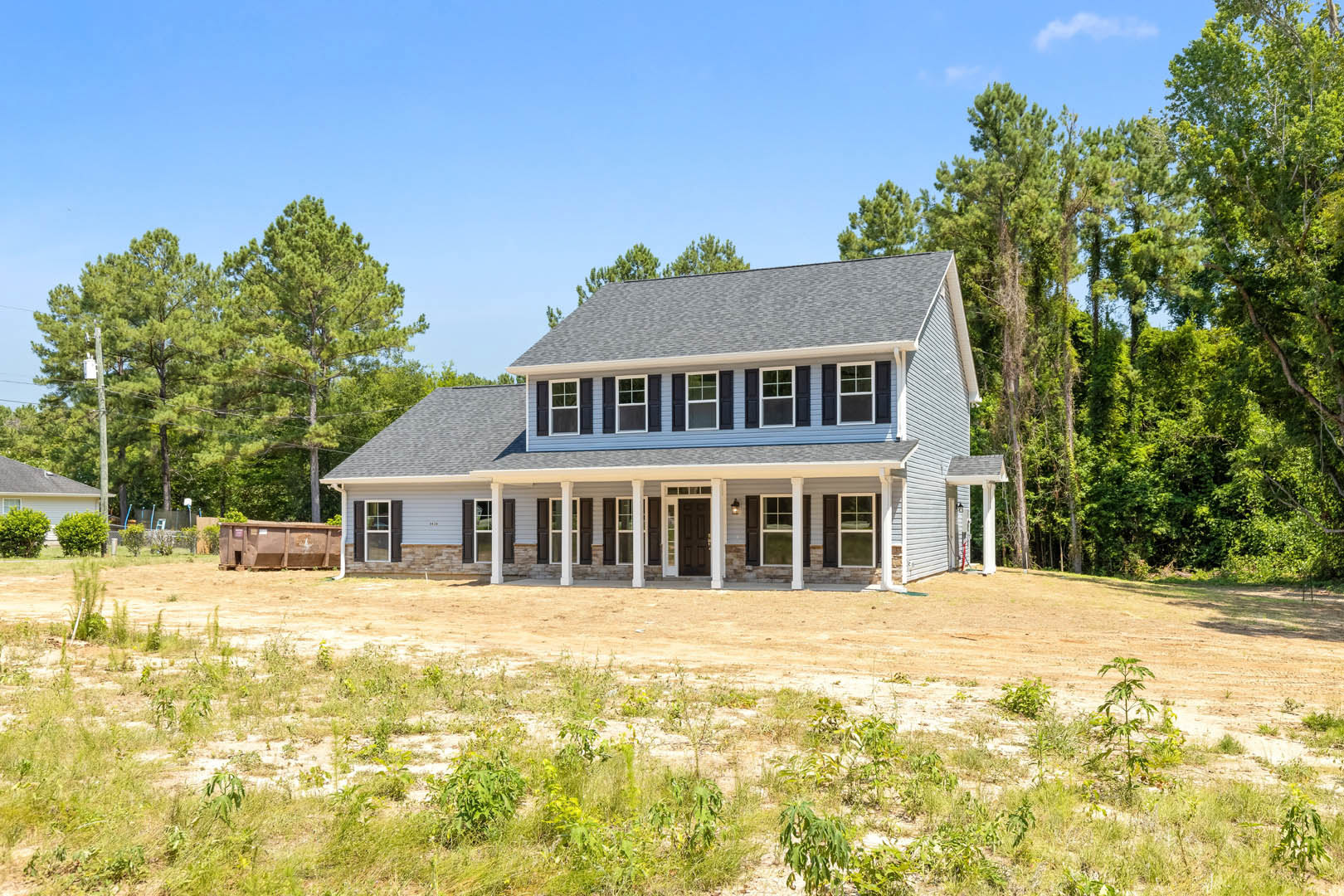 Large front porch with white columns, brown door, green bushes, and dirt field in foreground