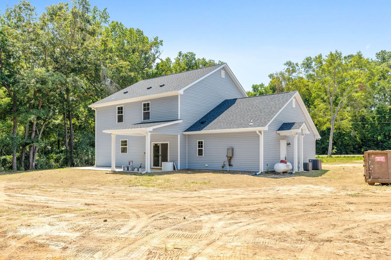 Partially built house with exposed framing, covered porch, glass entry door with side light, brown siding, dirt yard, white tank with red top, surrounded by trees.