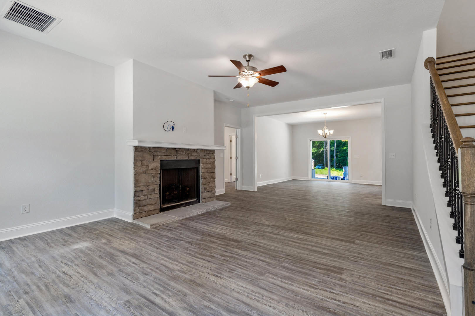 Living room with hardwood flooring, central fireplace with glass door, ceiling fan and light fixture, plaster walls, ceiling vent, large window showing outdoor view