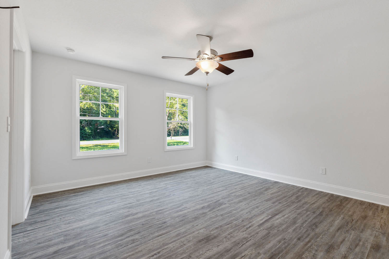 Ceiling fan with light fixture above wood flooring, large windows with views of trees, white plaster walls