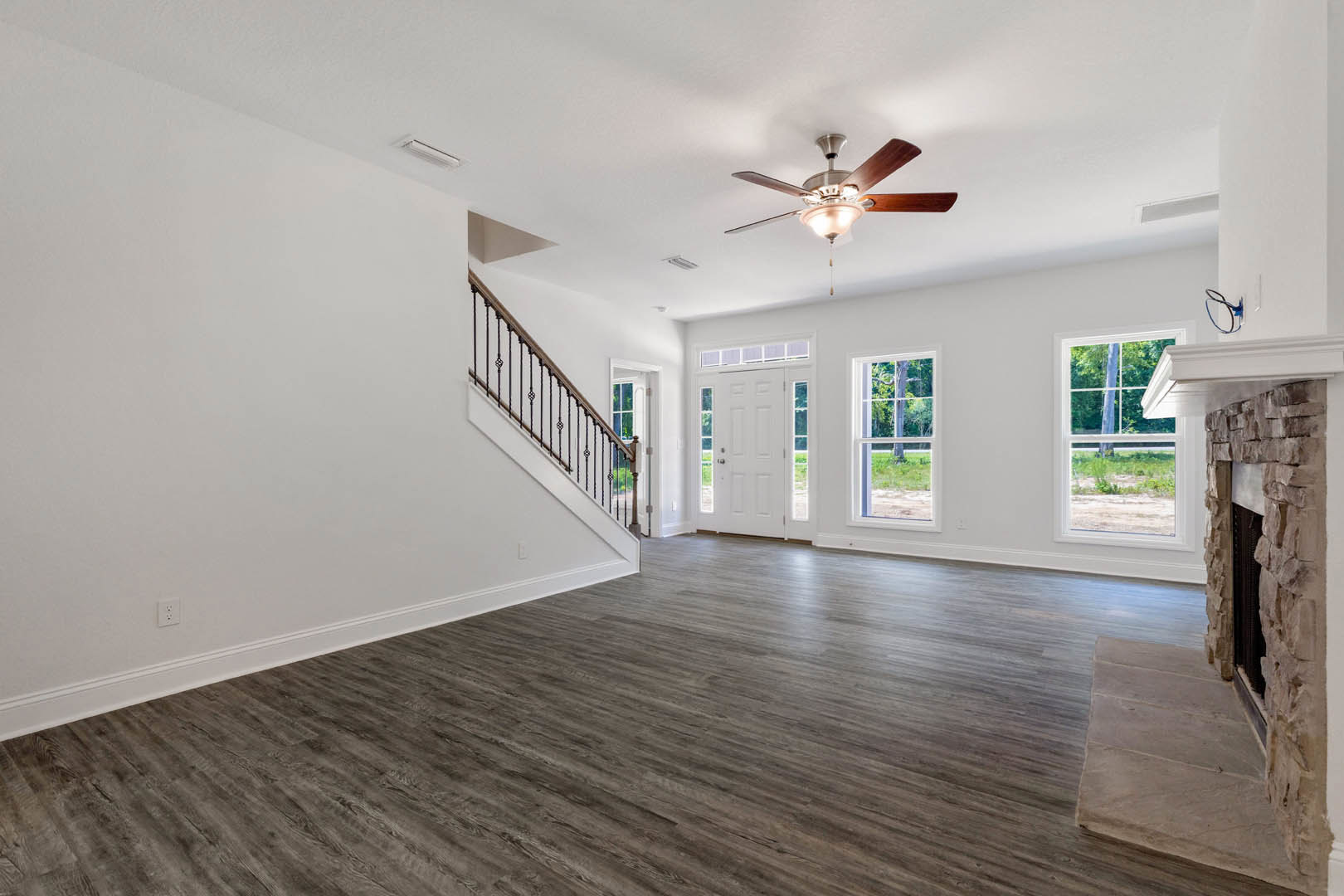 Hardwood floored room with white plaster walls, ceiling fan with light, wooden staircase, large window overlooking green lawn and tree, blue hoop resting on white surface